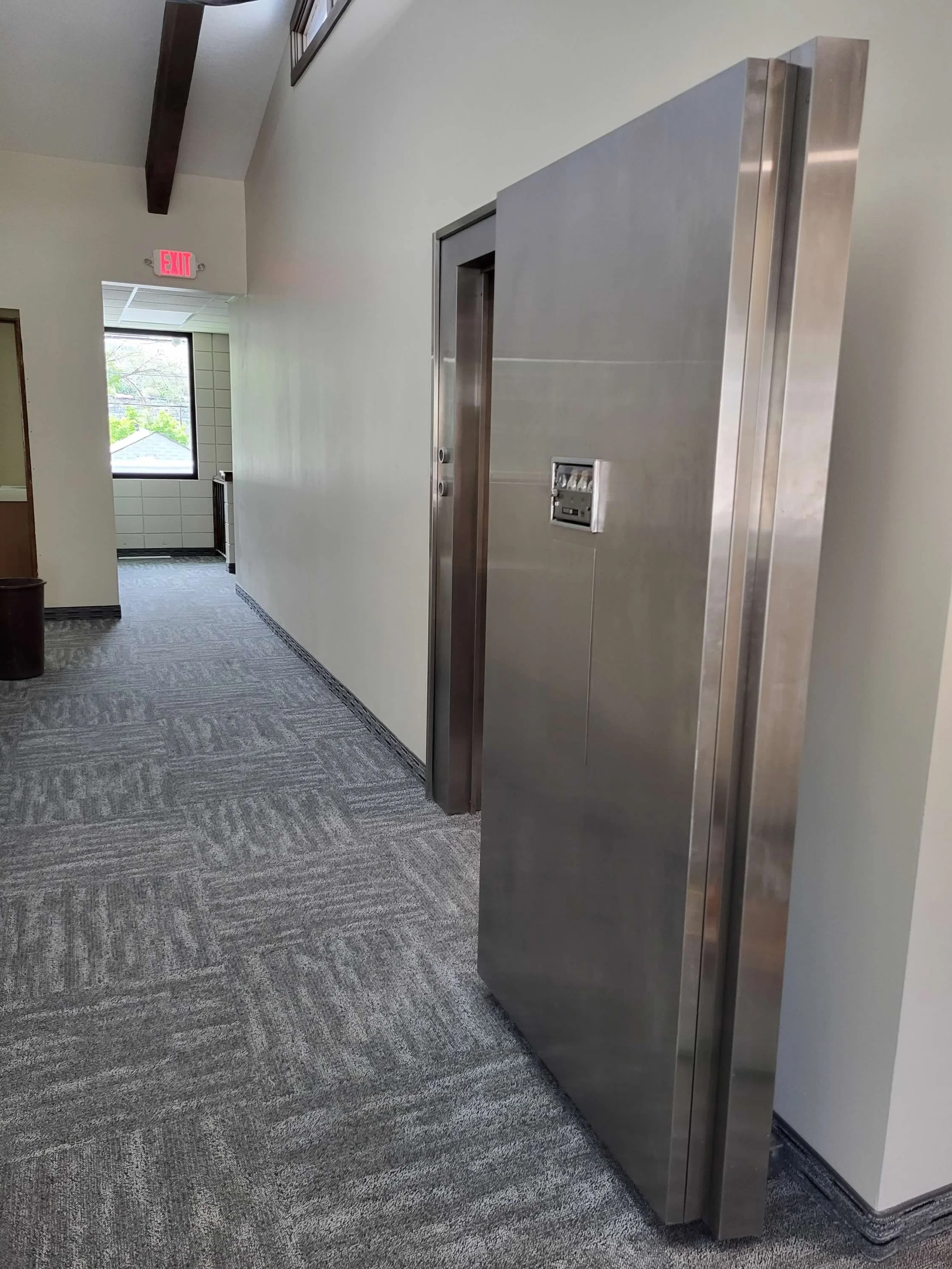 An elevator in a hallway with a stainless steel exterior, positioned against a white wall, with a control panel on the door. The hallway has a gray patterned carpet, a window at the end, and an EXIT sign overhead.