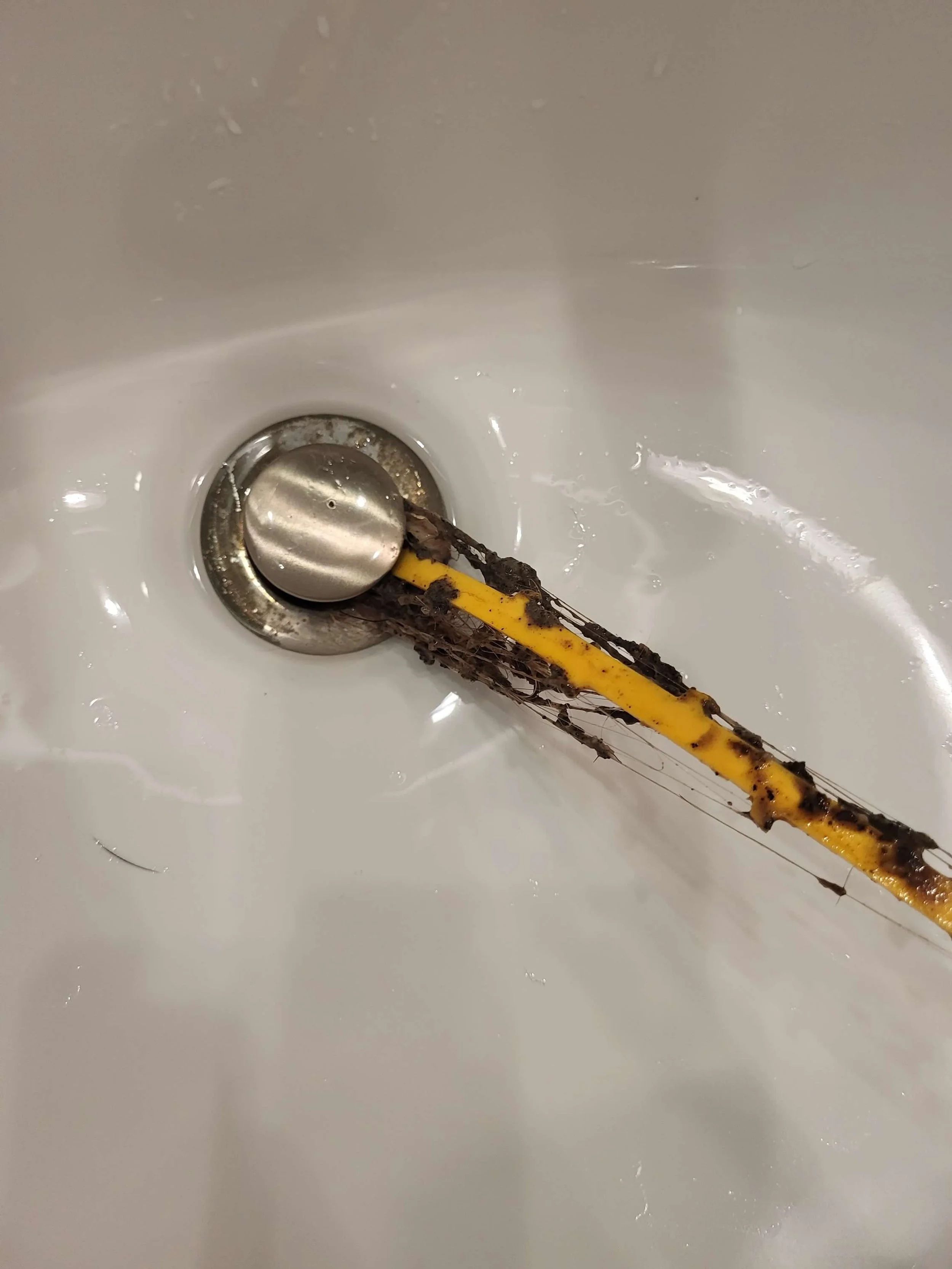 A yellow and gray cleaning brush with black bristles and dirt on it, resting in a white sink drain.