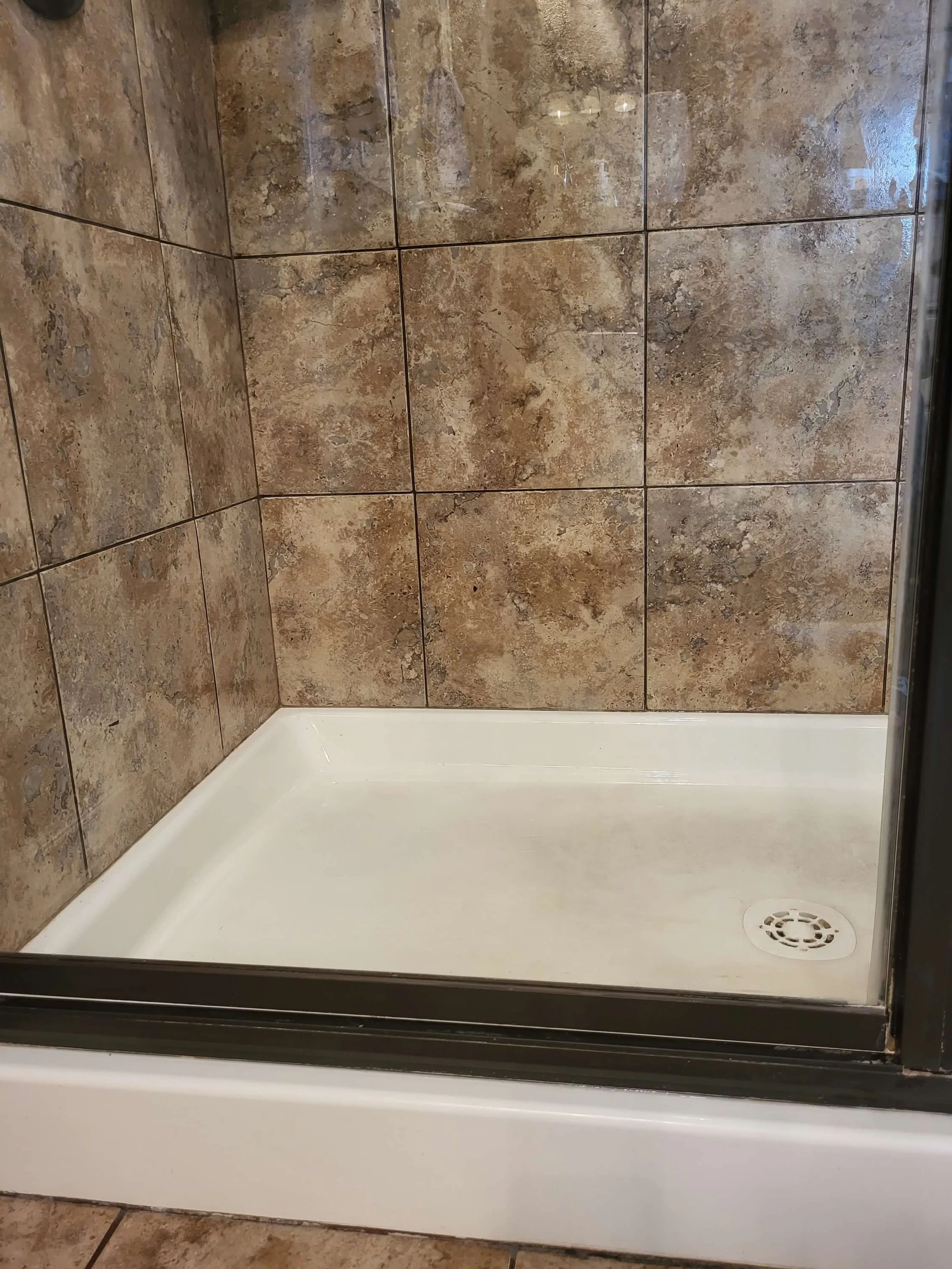 Empty shower stall with beige and brown stone tile walls, white shower pan, and a round drain in the corner.