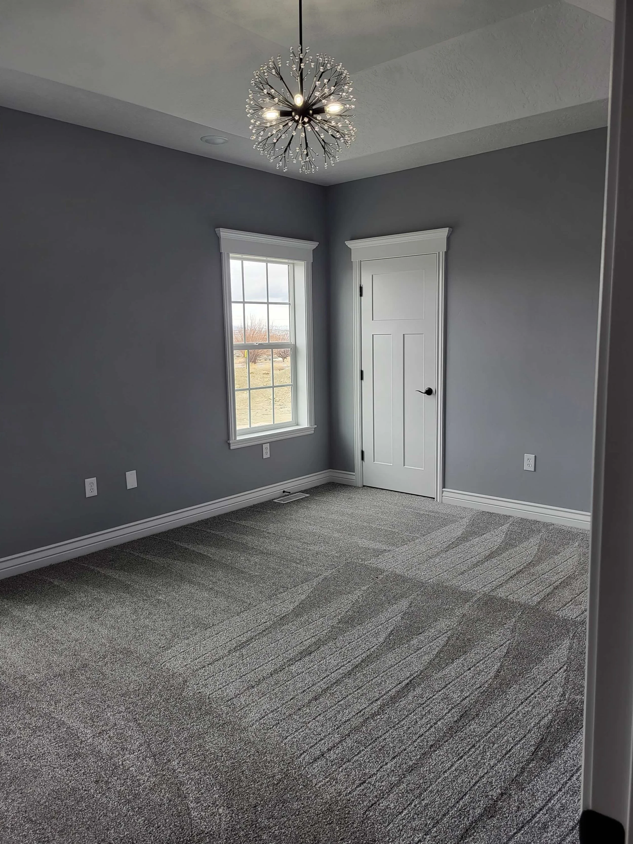 Empty bedroom with gray walls, white trim, gray carpet, a window, a white door, and a modern chandelier.