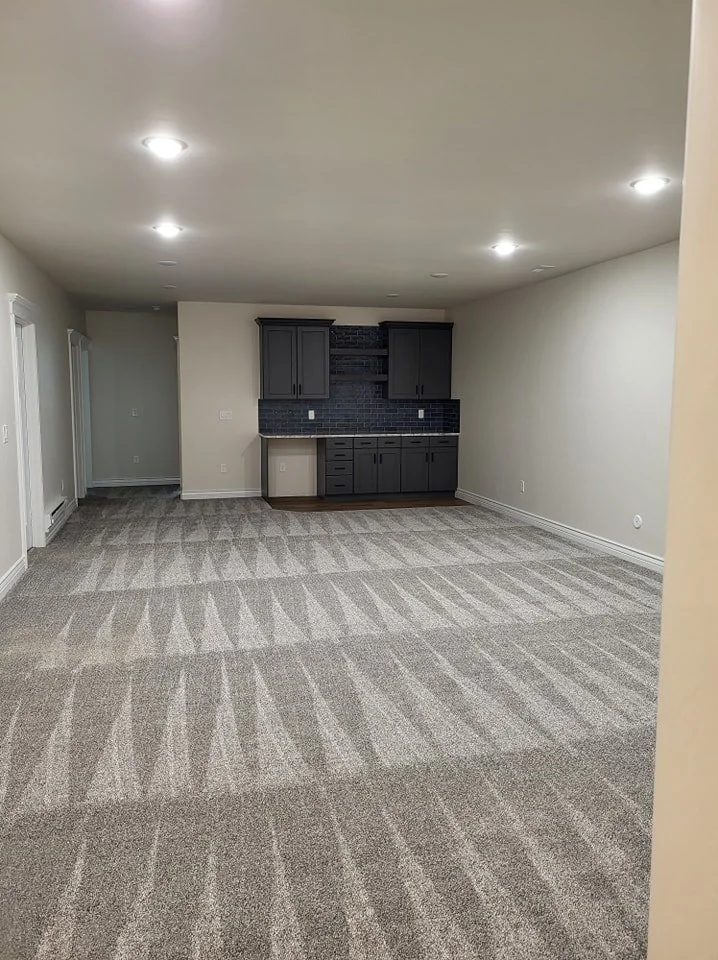 Empty living room with carpeted floor, off-white walls, and ceiling lights, featuring a small dark gray kitchenette with black backsplash.