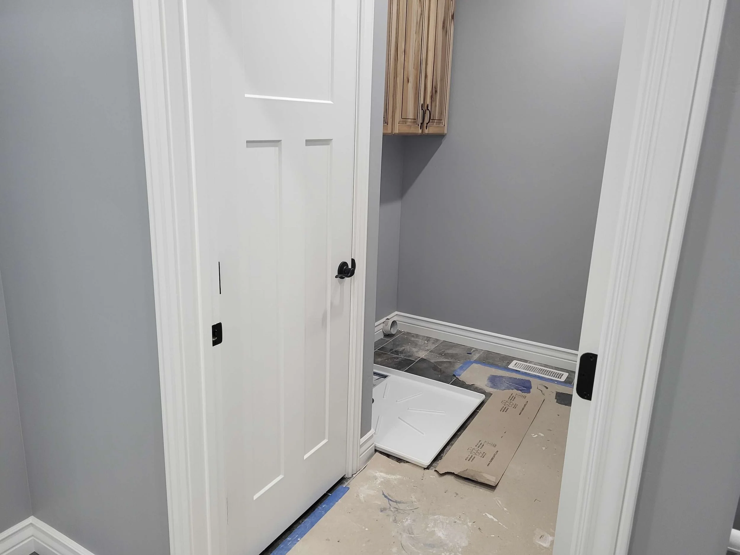 Interior view of a laundry or utility room with a closet door, gray walls, wooden cabinets, partially installed flooring, and a wash basin or utility sink.