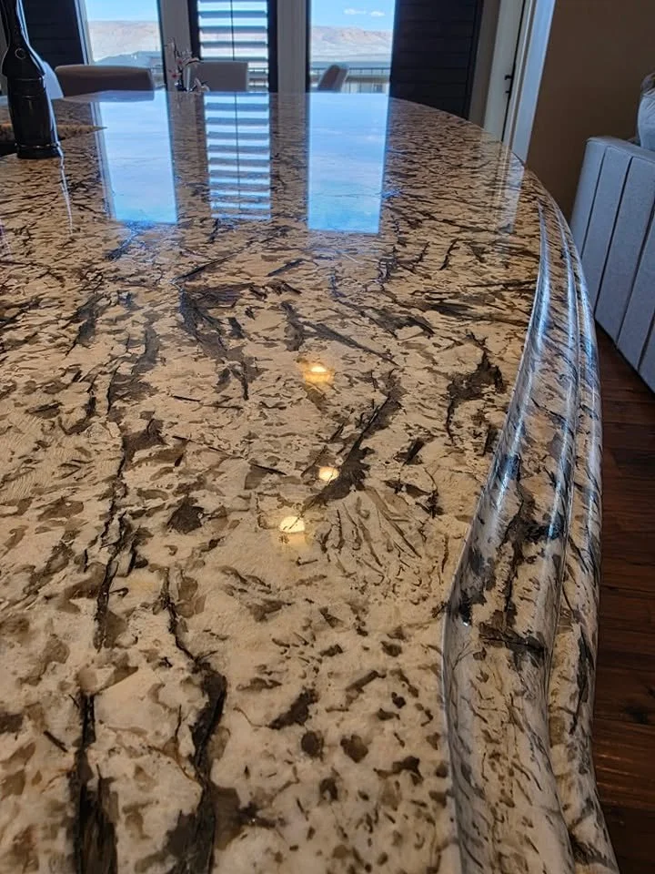 Close-up of a polished granite countertop with a beige and black marbled pattern, reflecting overhead lights in a modern kitchen or dining area.