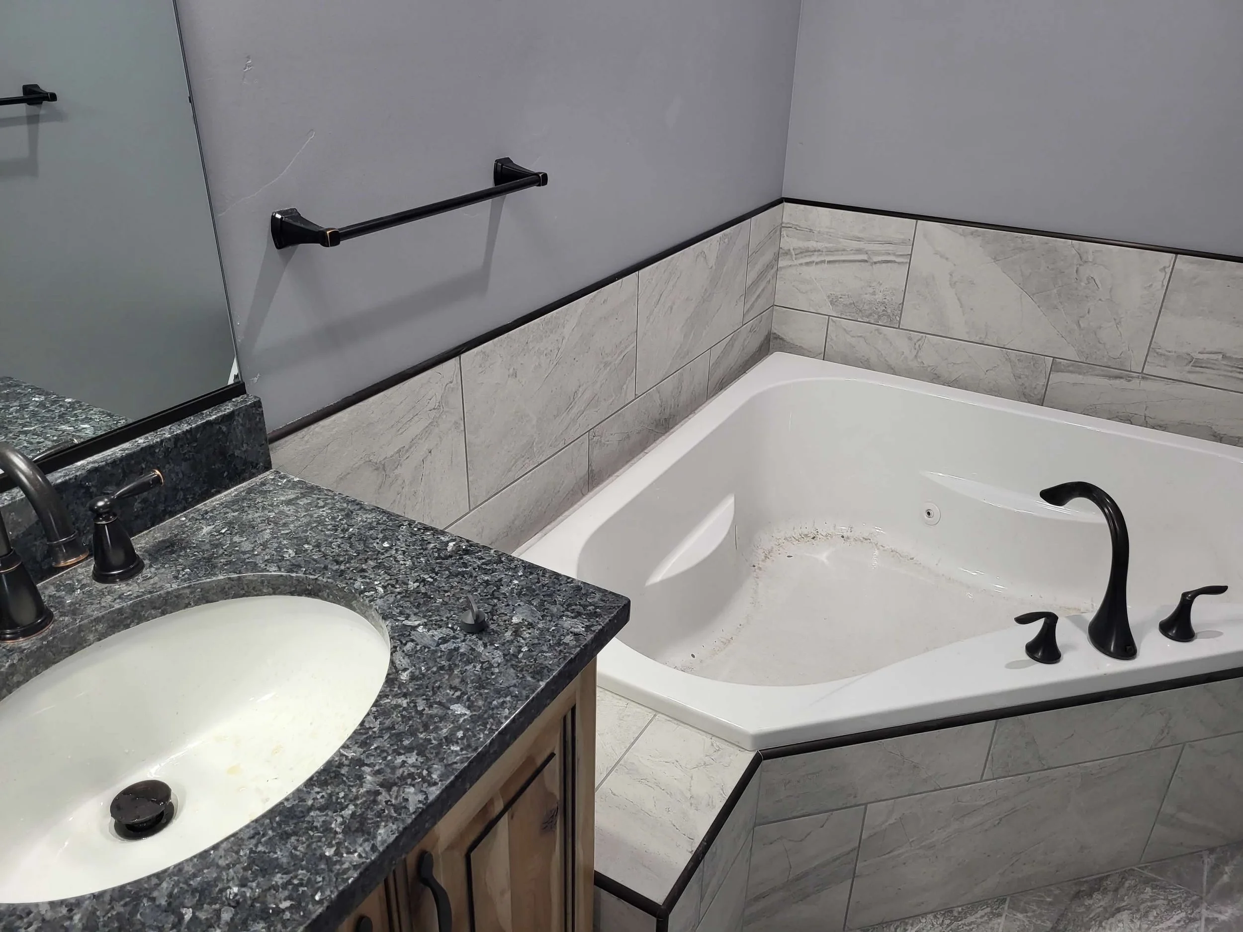 Bathroom with a granite countertop vanity, a sink, a mirror, a gray towel bar, a corner bathtub with black fixtures, and marble wall tiles.