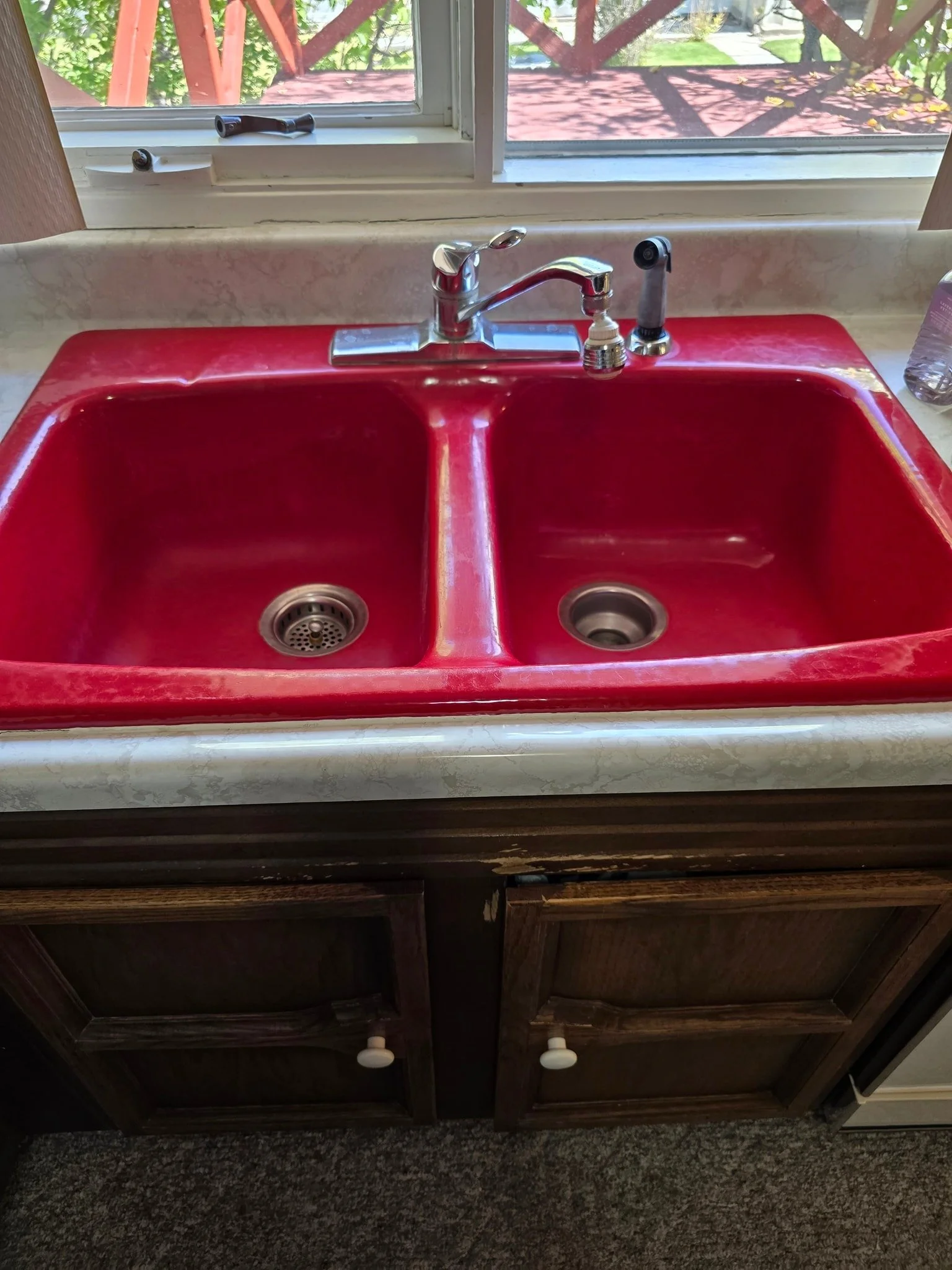 A red double-basin kitchen sink with a chrome faucet, positioned below a window overlooking a backyard with a red deck.