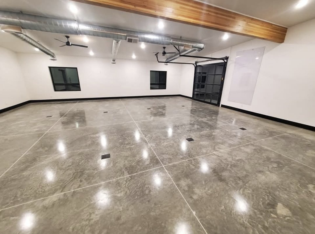Empty industrial-style garage with polished concrete floors, white walls, two small windows, ductwork on the ceiling, black trim, a garage door, and ceiling fans.