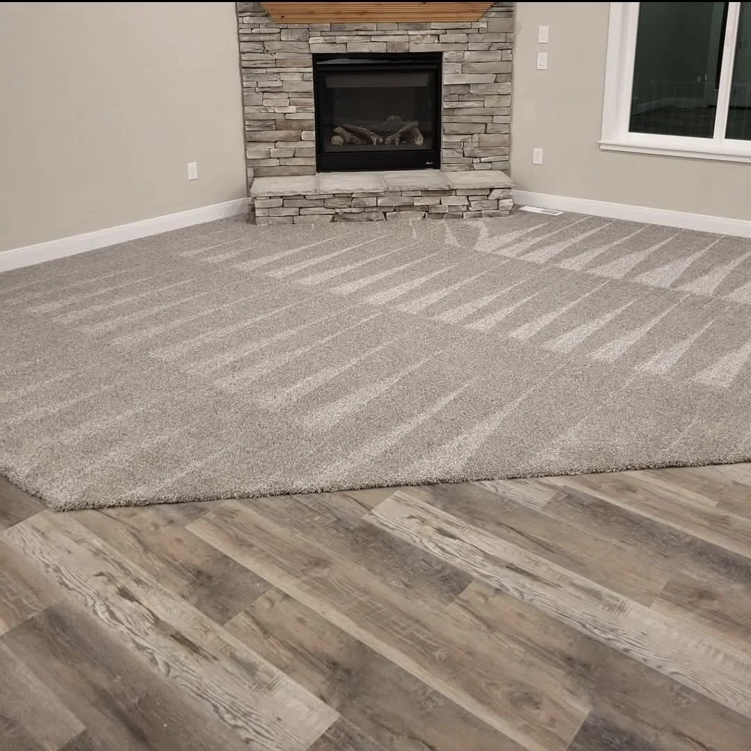 A living room with a fireplace, beige walls, a gray carpet, and wood-look flooring. The carpet has vacuum lines showing progress, and there is a window on the right side.