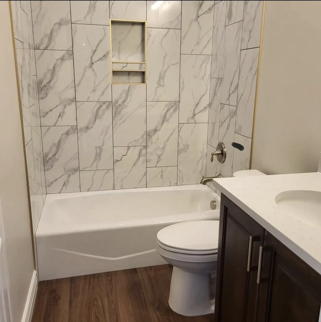 A bathroom with a marble-tiled shower wall, a white bathtub, a toilet, and a dark cabinet with a white countertop and sink.