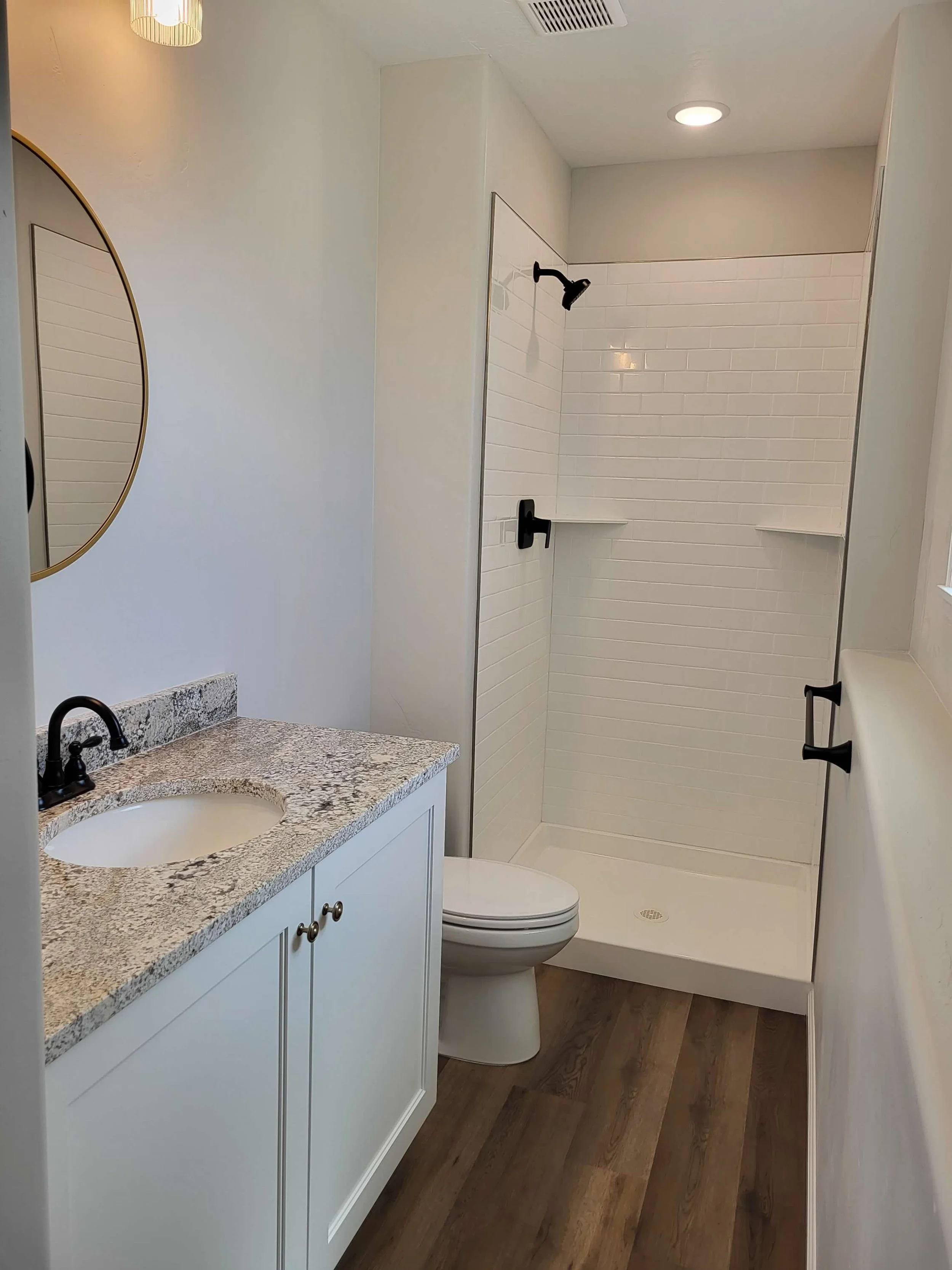 A small bathroom with a white vanity, granite countertop, oval sink, black faucet, and round mirror. Next to it is a toilet and a walk-in shower with white subway tiles, black fixtures, and built-in shelves. The floor is wood.
