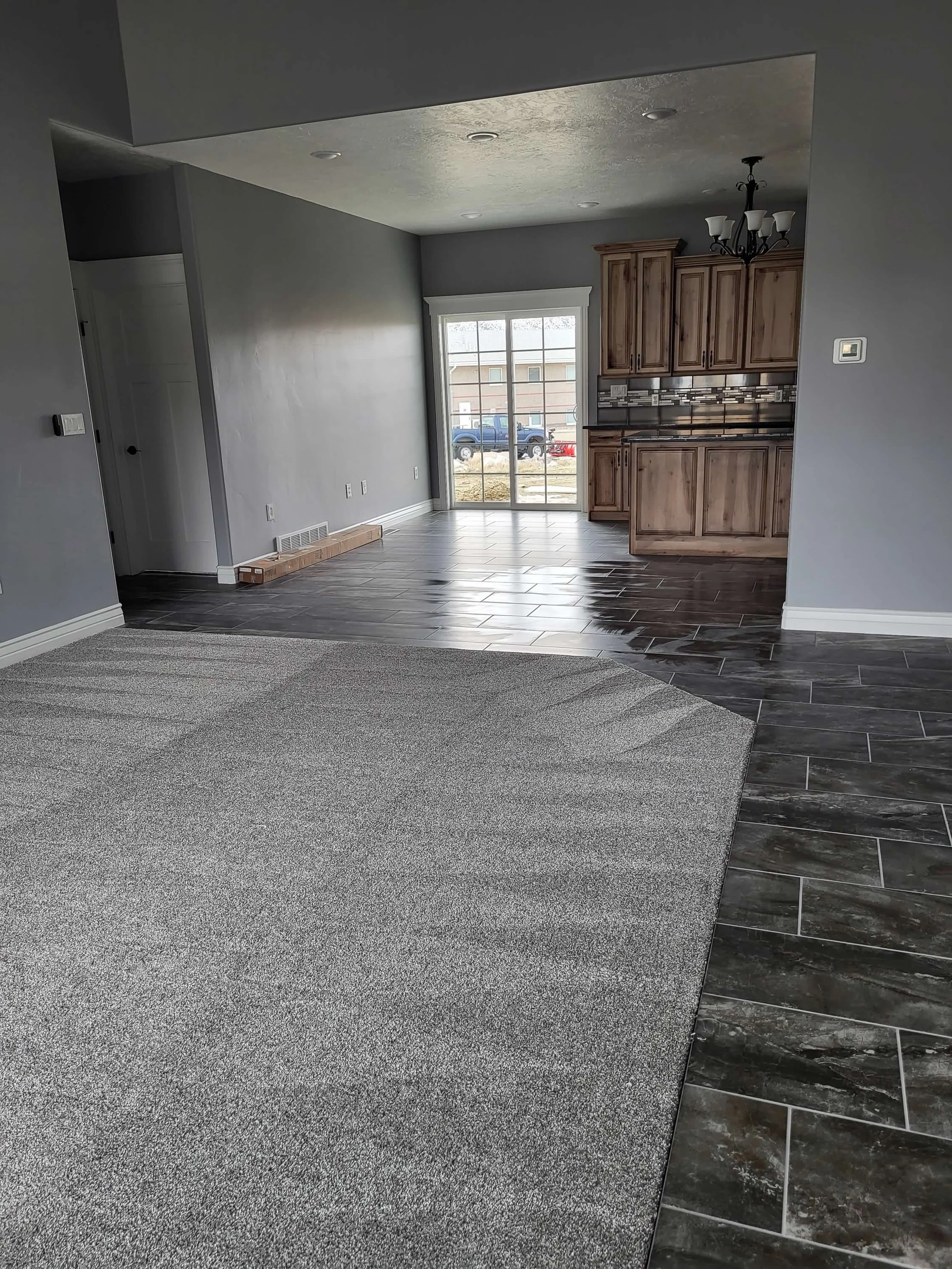 Empty living room and kitchen with gray walls, tile floors, and wooden cabinets, illuminated by natural light from a sliding glass door.