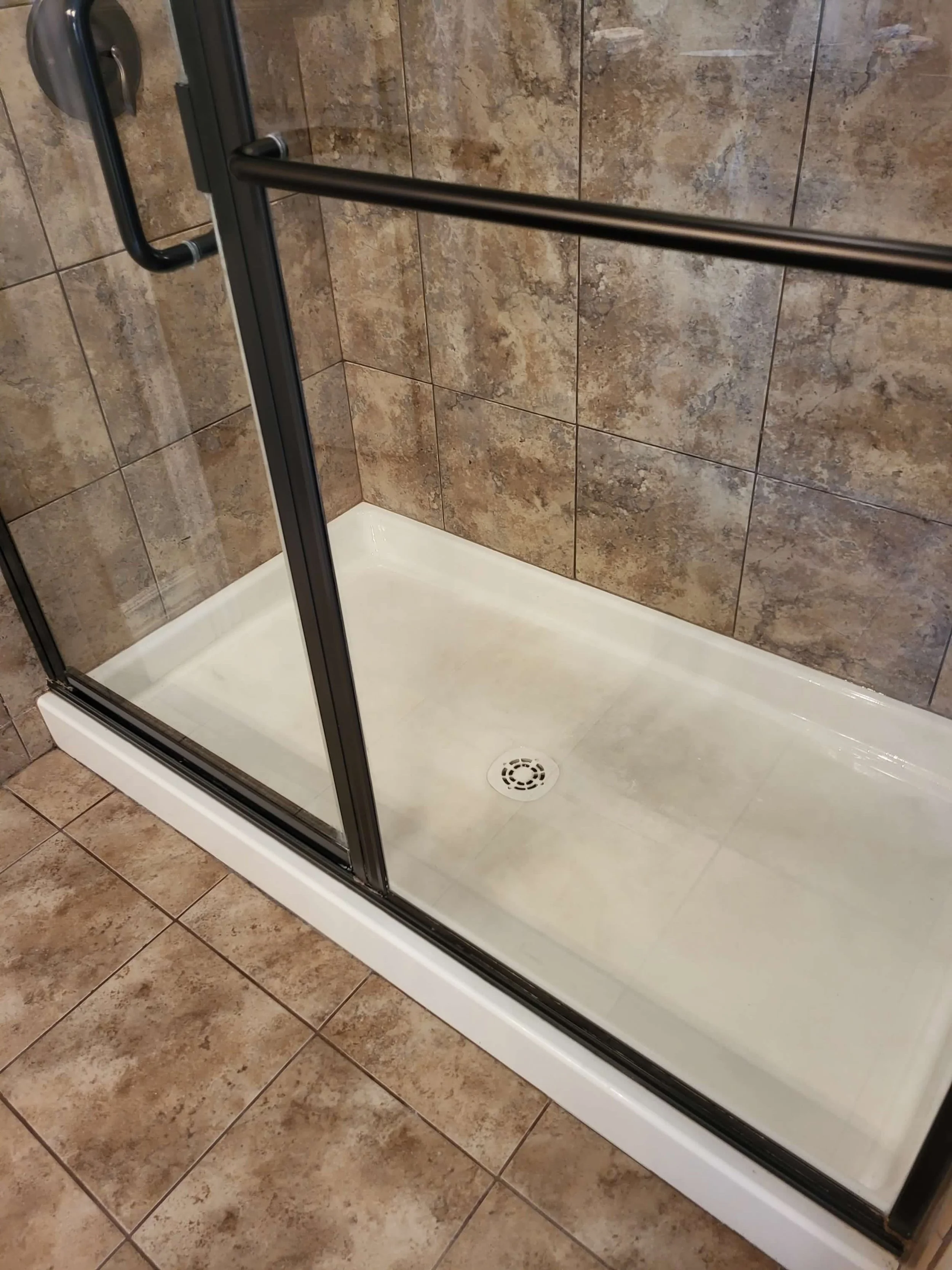 A glass shower with a sliding door, tan tiled walls and floor, and a white shower base with a drain in the center.