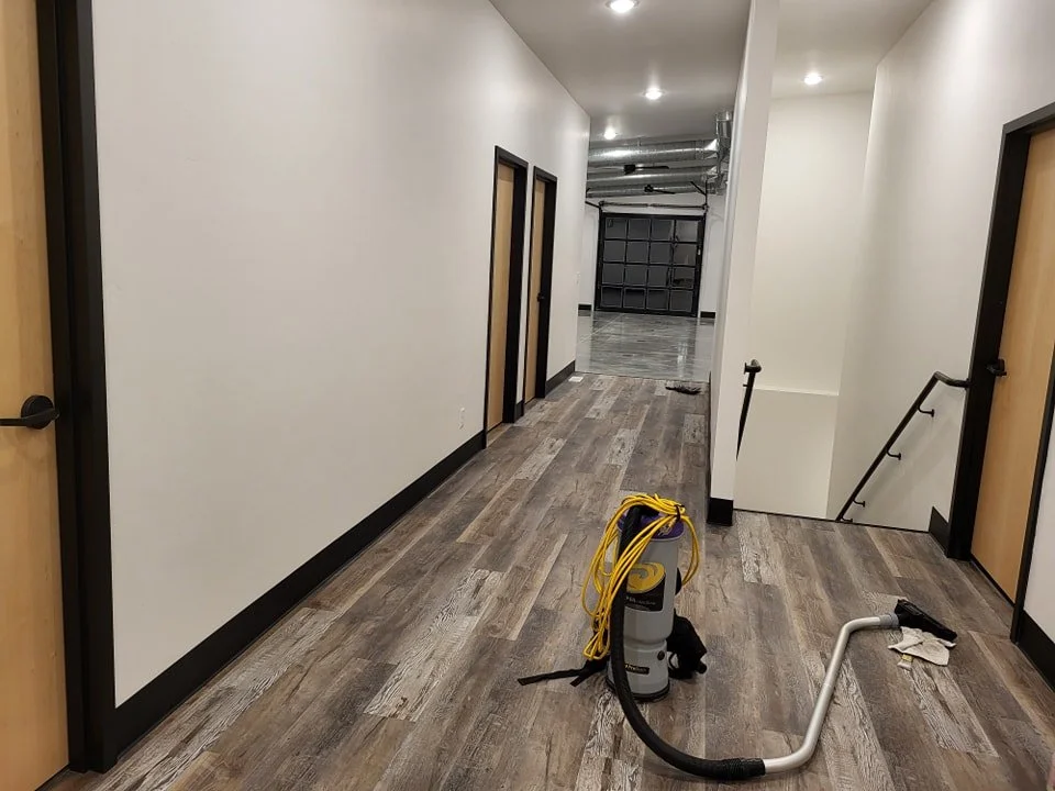 Inside a modern building hallway with light-colored walls and wood-look flooring. There are closed doors on the left side and an open area with a garage-style door at the end. A vacuum cleaner and cleaning tools are in the foreground.
