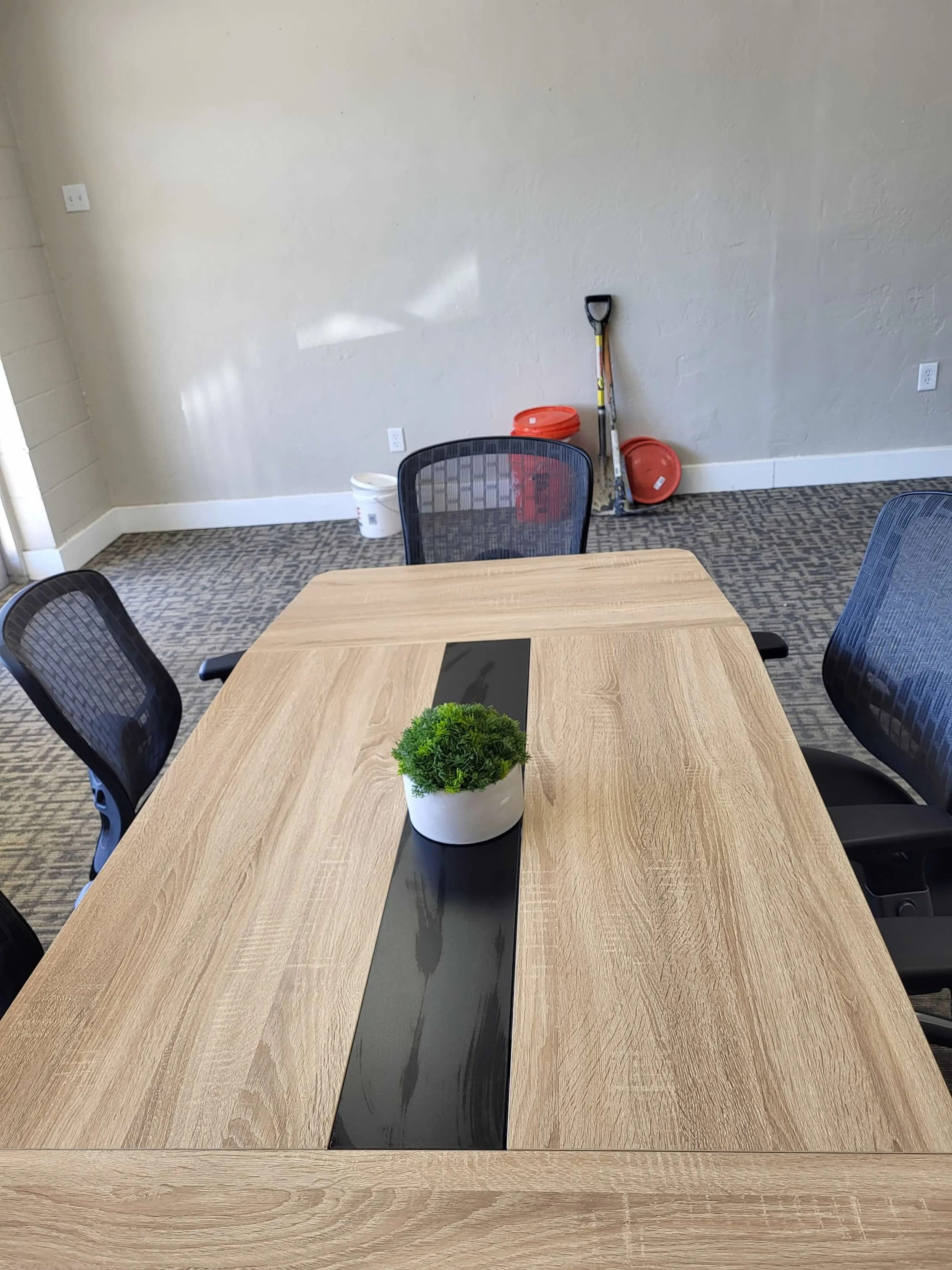 Empty conference room table with a small potted green plant in the center, surrounded by black office chairs. In the background, there are cleaning supplies and tools against the wall.