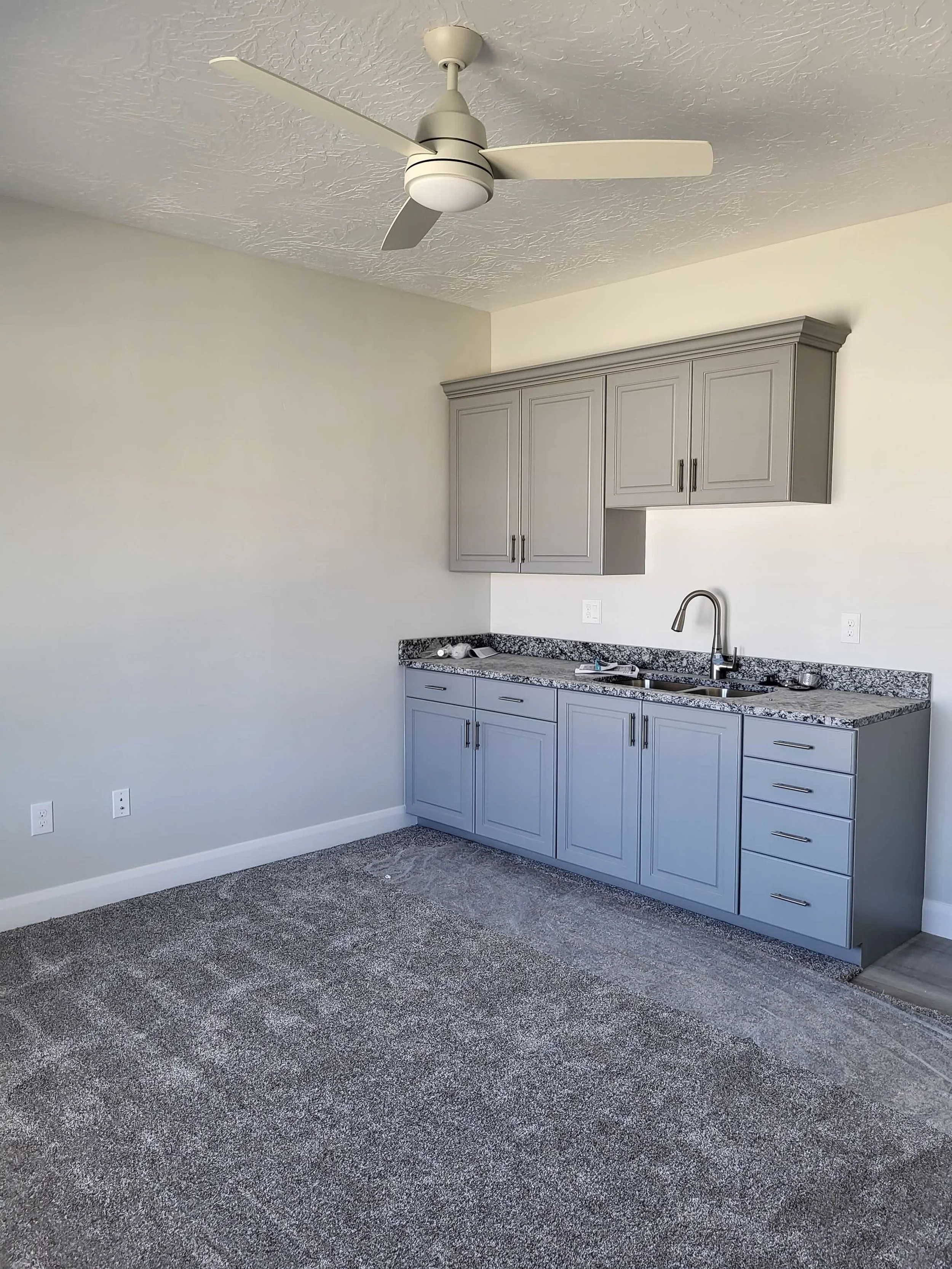 Empty kitchen with gray cabinets, granite countertop, white walls, ceiling fan, and gray carpet.