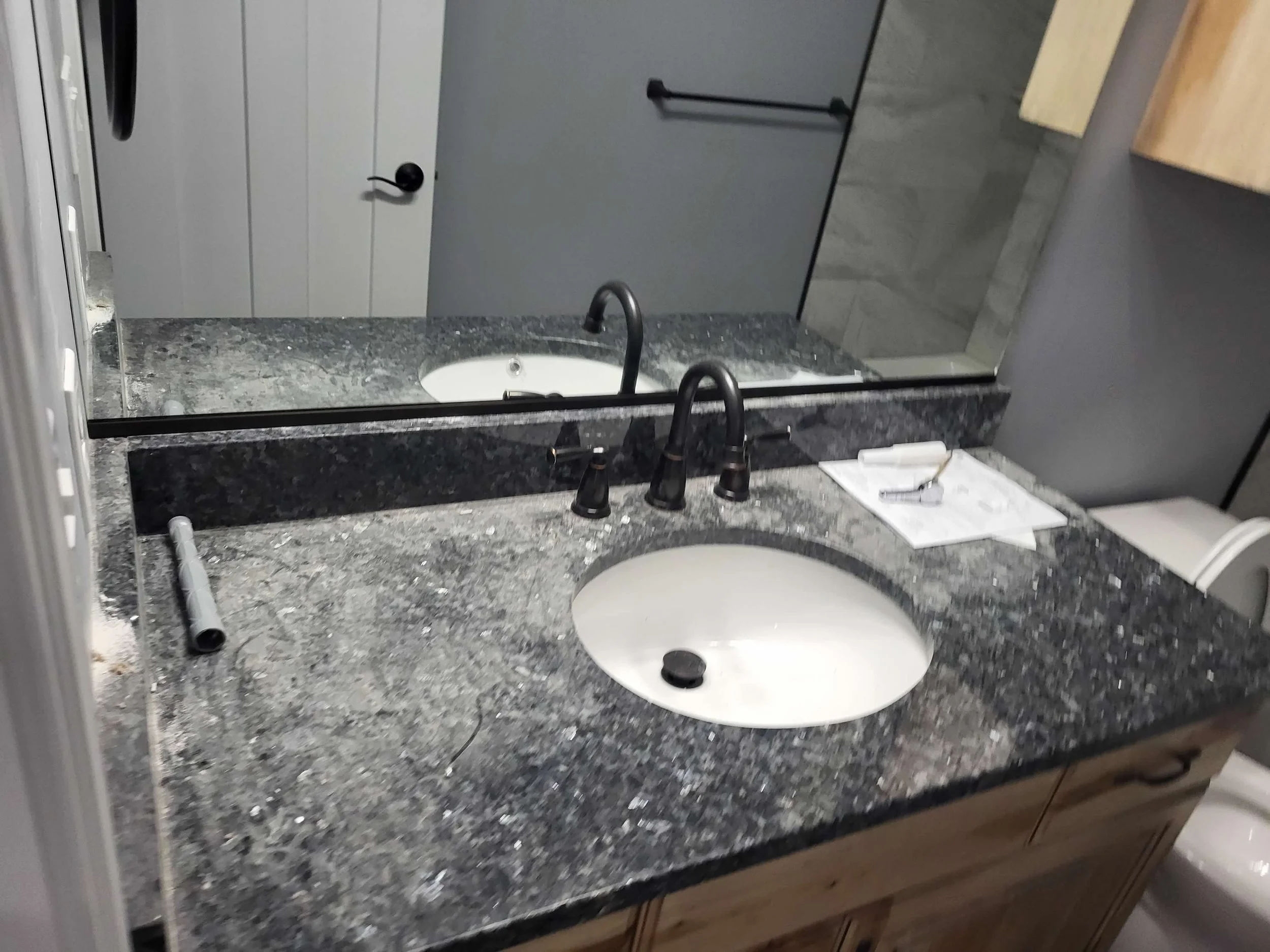 Bathroom vanity with a granite countertop, white oval sink, black faucet, mirror, tools, and papers on the counter.