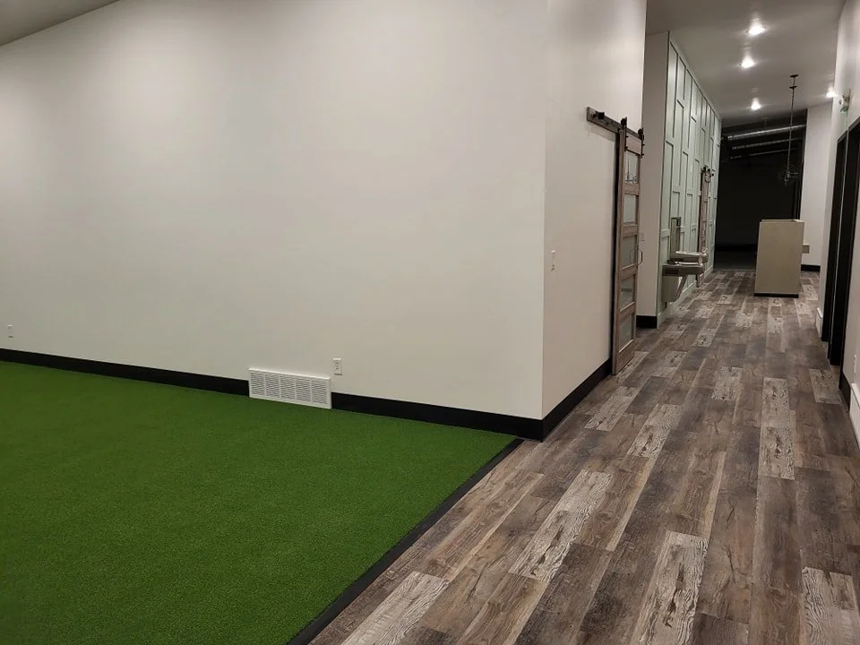 Empty indoor corridor with a small green artificial turf area on the left, white walls, wood-style flooring, and a water fountain along the right wall.