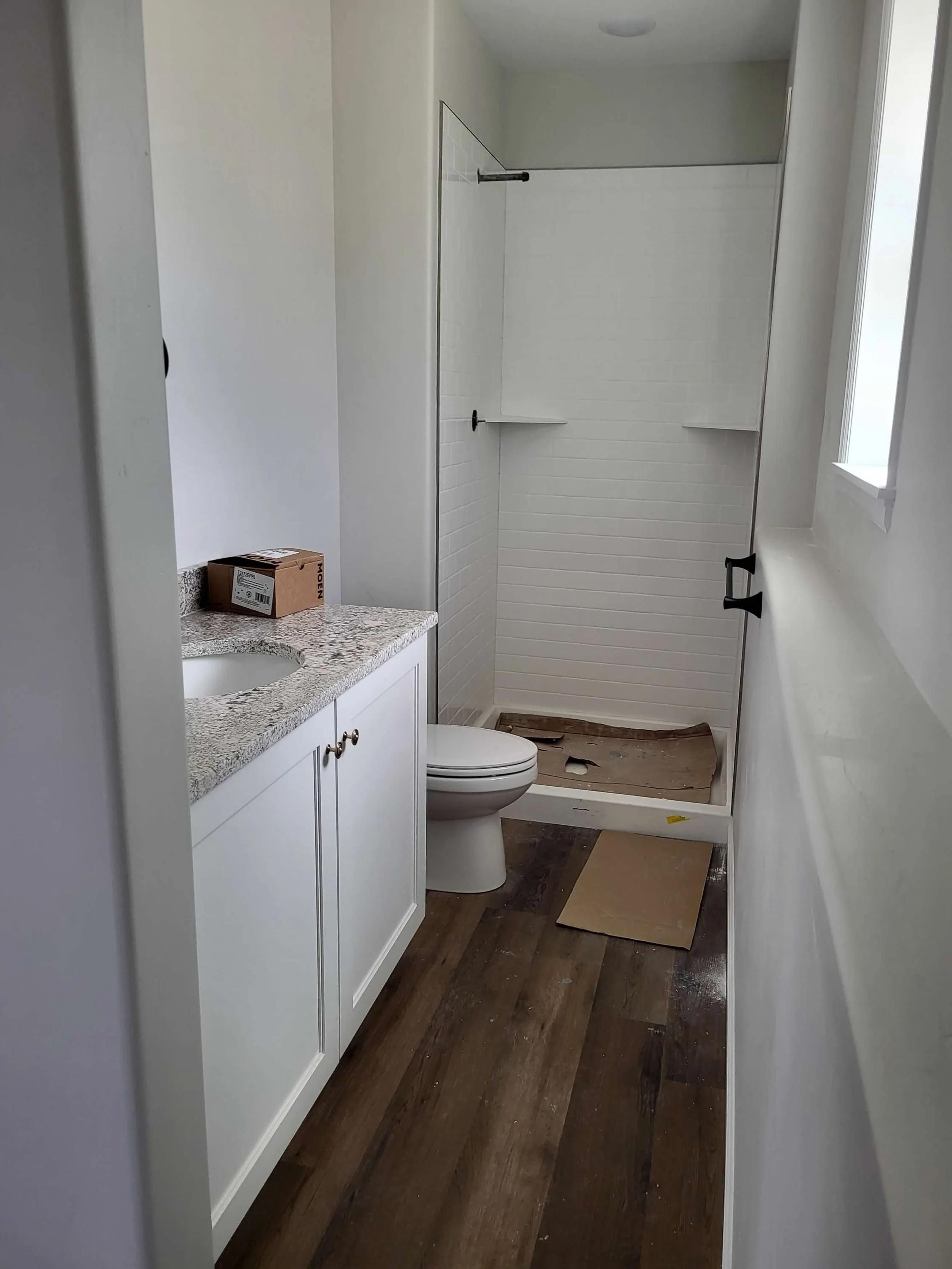 Bathroom with white vanity, granite countertop, toilet, and a shower with a black bar, white tile walls, and built-in shelves. There is a small window and a brown papered floor, possibly under renovation.