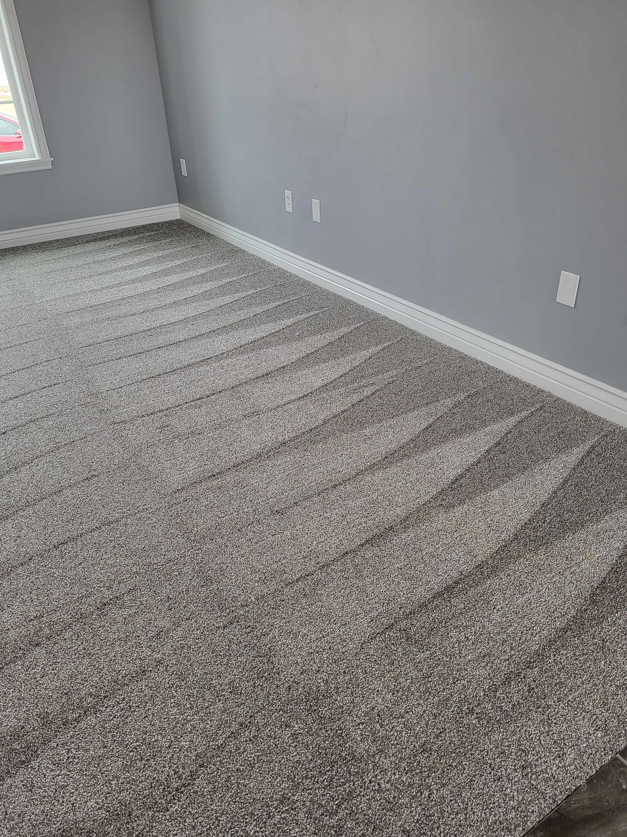 Empty room with gray walls, white baseboards, carpeted floor with vacuum lines, and a window on the left side.