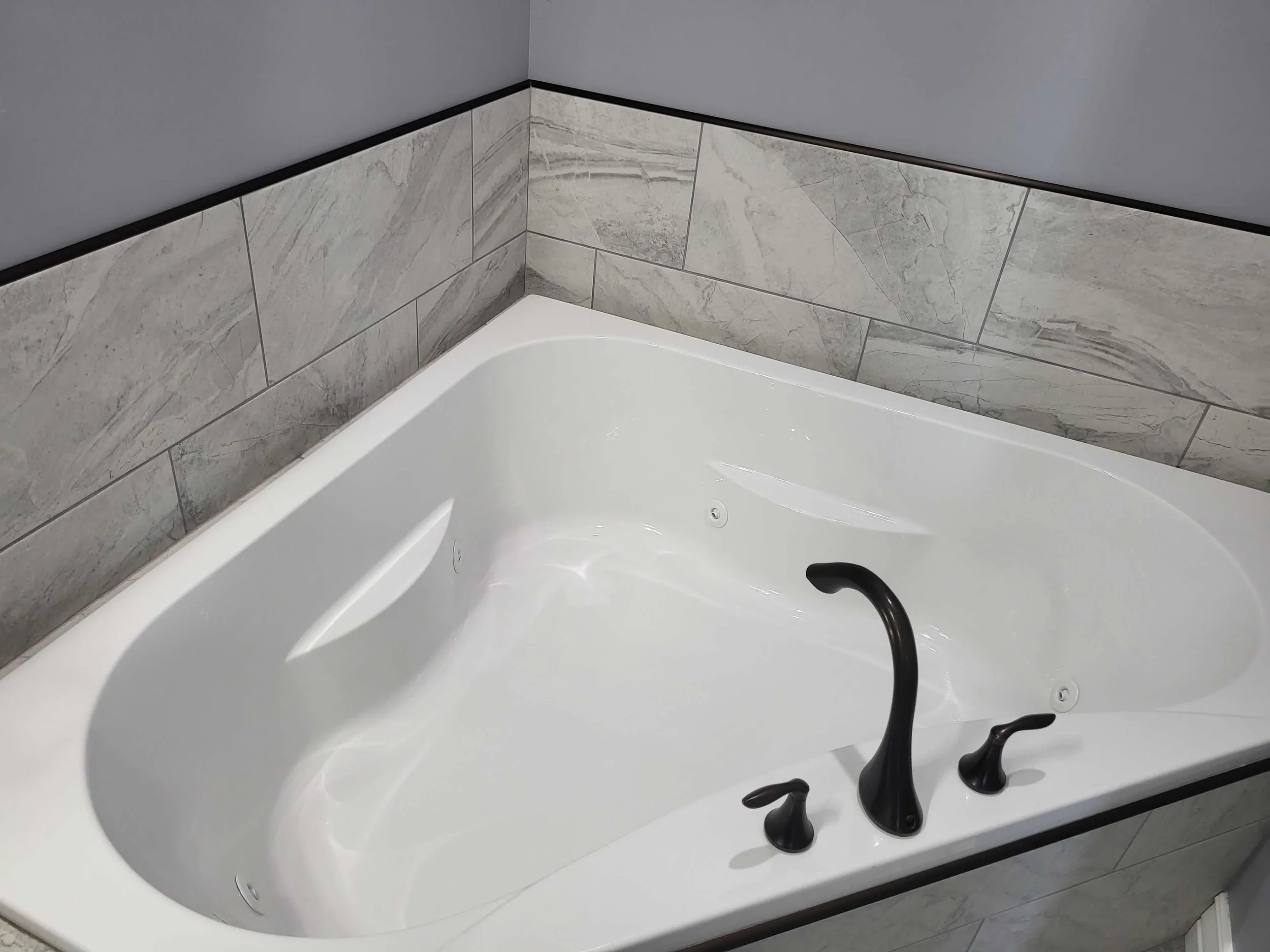 Empty white bathtub with black fixtures, set against a tiled wall with gray and white marble patterns.