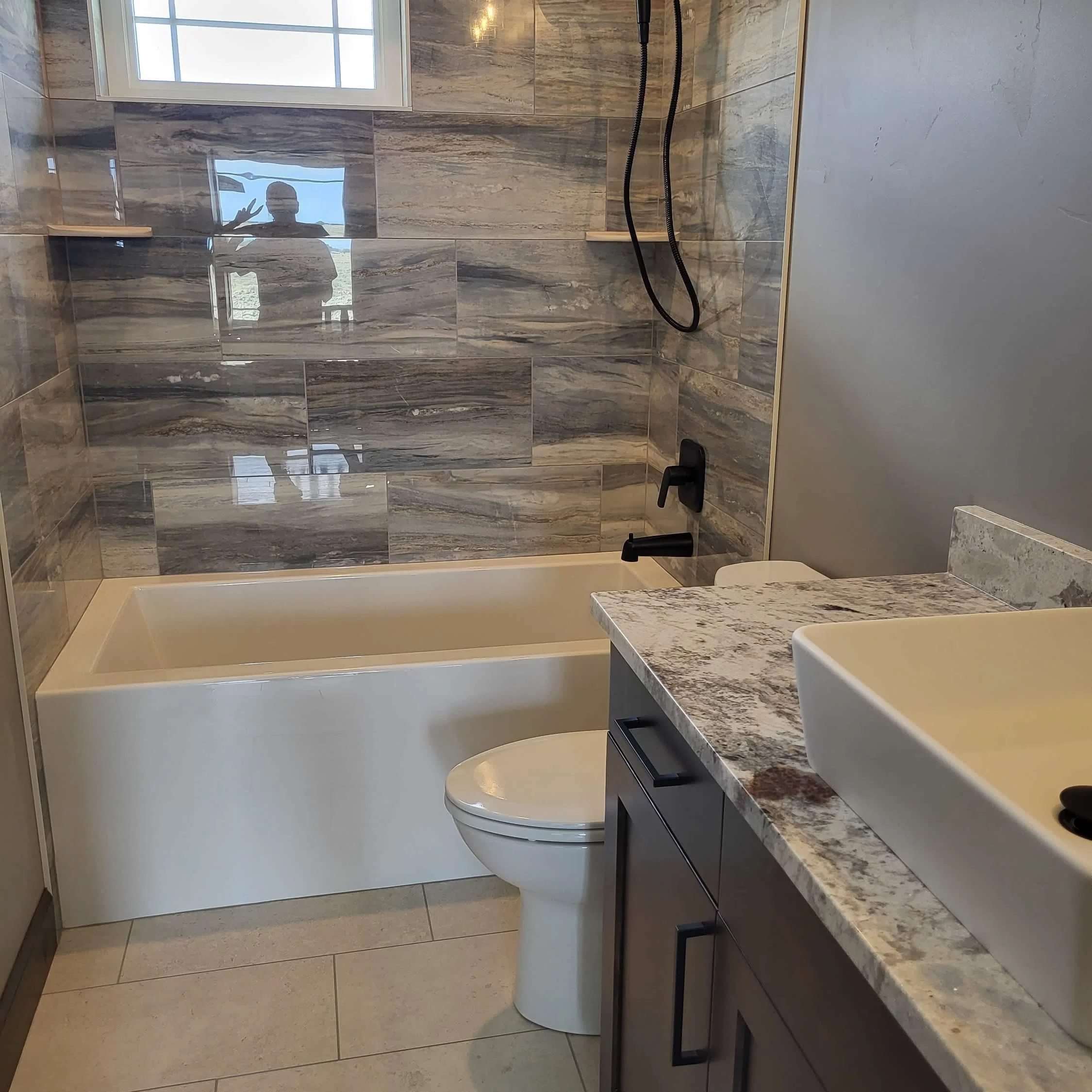 Modern bathroom with a bathtub, toilet, marble countertop with a vessel sink, and tiled walls with a window.