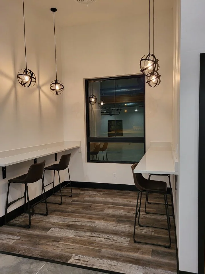 Empty corner with two wall-mounted white counters and three chairs, illuminated by pendant lights, with a window showing an empty room with additional chairs.