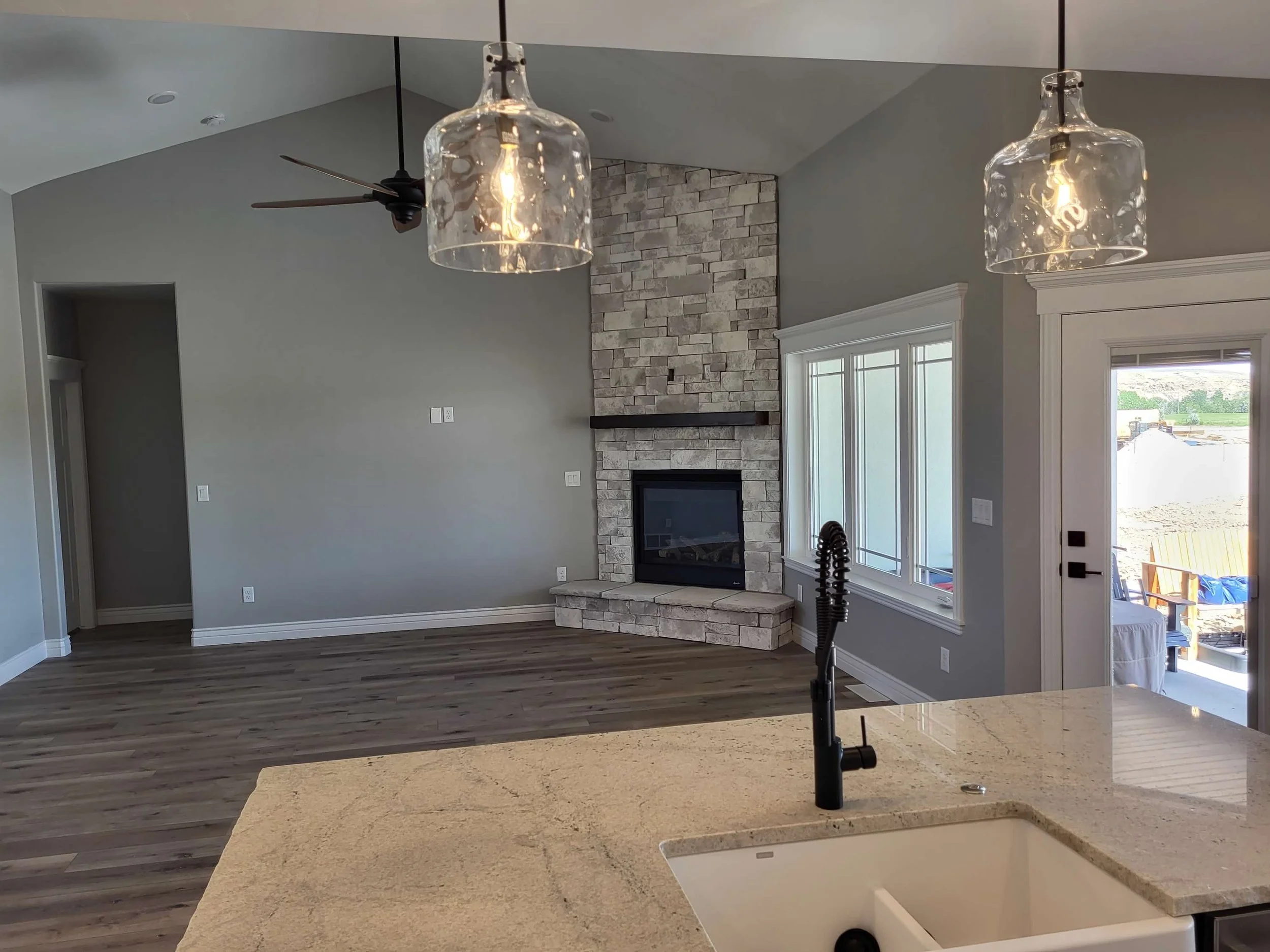 Open-concept living room with stone fireplace, hardwood flooring, large windows, and a screened porch outside. Two glass pendant lights hang above a kitchen island with a modern black faucet.