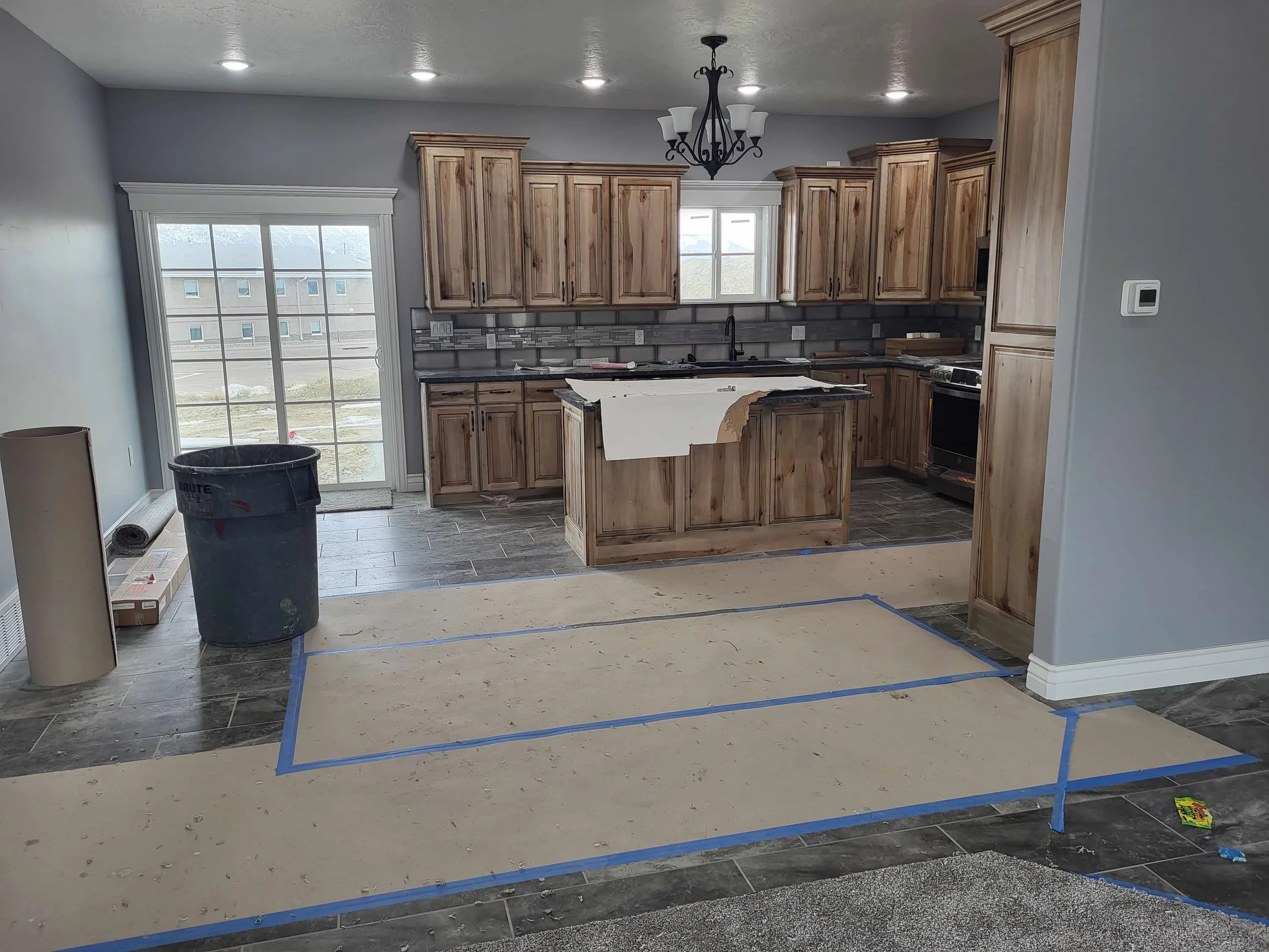Kitchen under construction with unfinished flooring, wooden cabinets, gray tile backsplash, and a black chandelier. A large sliding glass door leads outside. Construction materials and tools are present.