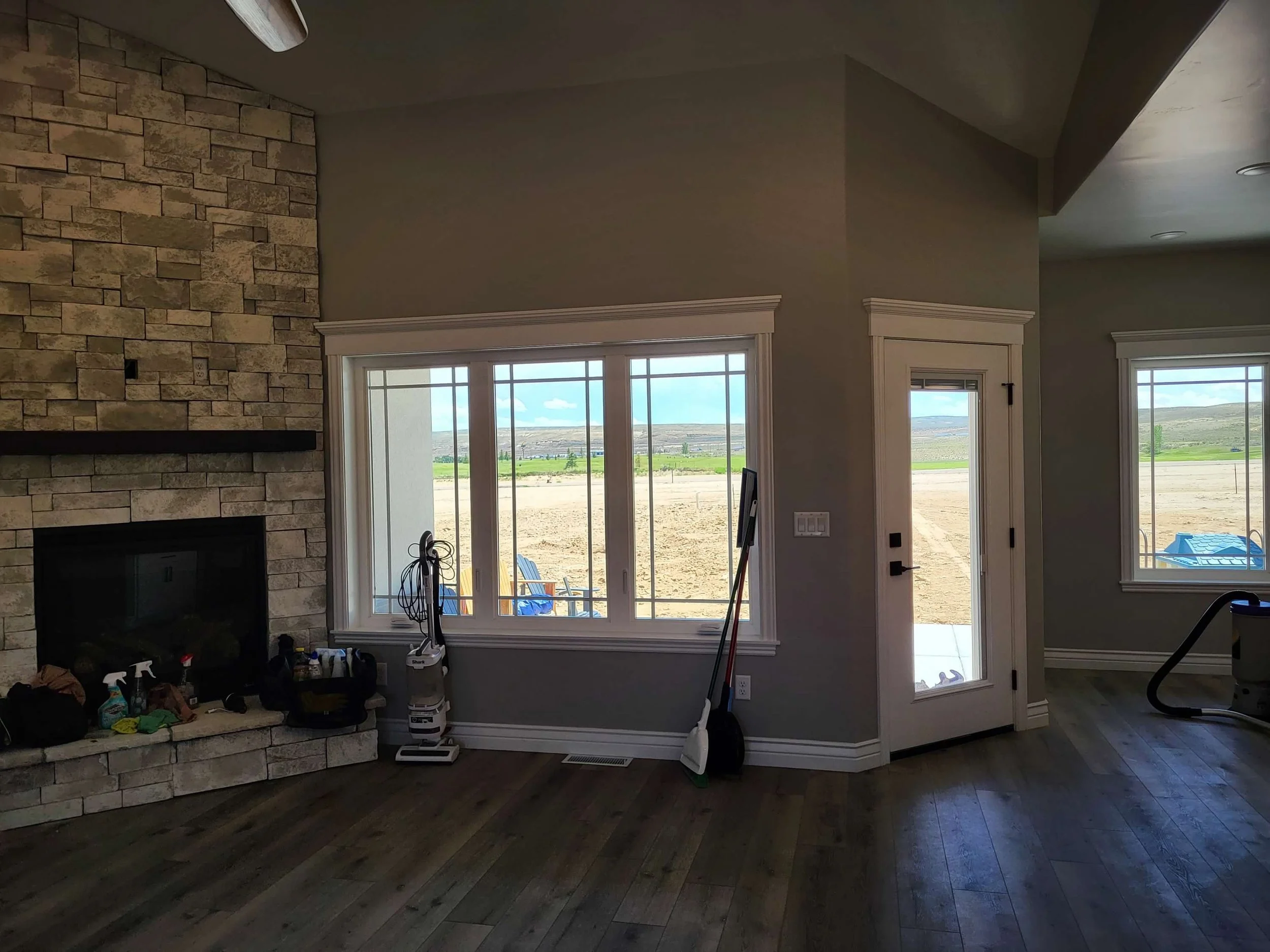 Interior view of a living room with large windows showing a landscape of dirt and distant green hills, a stone fireplace on the left, cleaning tools and supplies near the window, and hardwood floors.