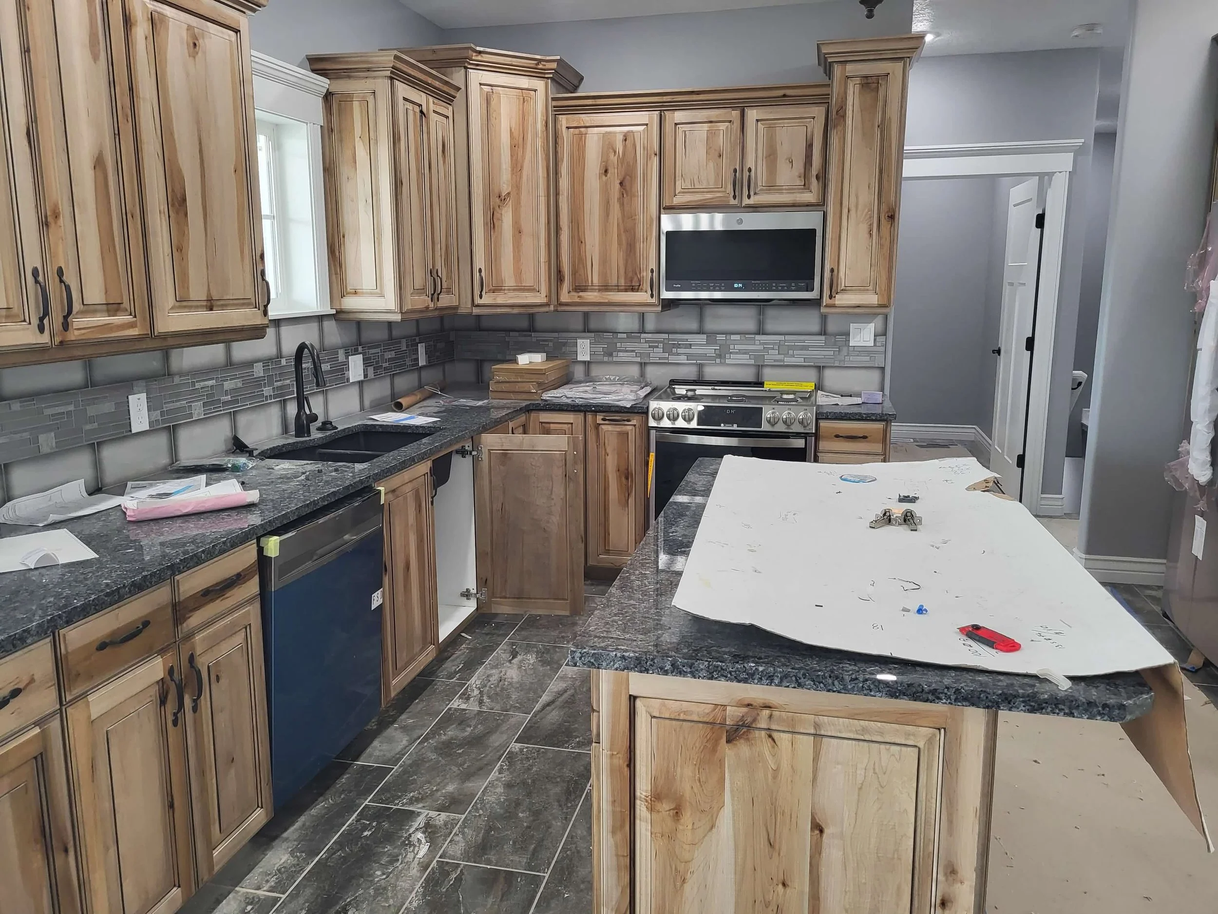 Kitchen with wooden cabinets, a black granite countertop, a stove, microwave, and a dishwasher. The kitchen is under renovation, with tools and papers on the counter and an island covered with white paper, construction items, and scattered objects.