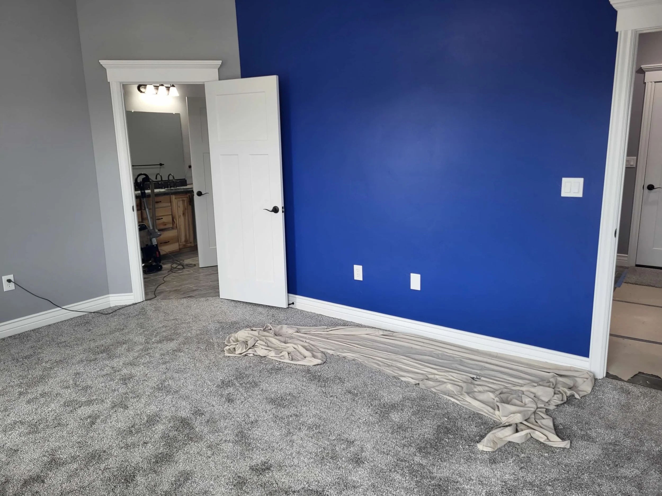 Empty room with gray carpeting, a blue accent wall, a white open door leading to a bathroom, and a beige drop cloth on the floor.