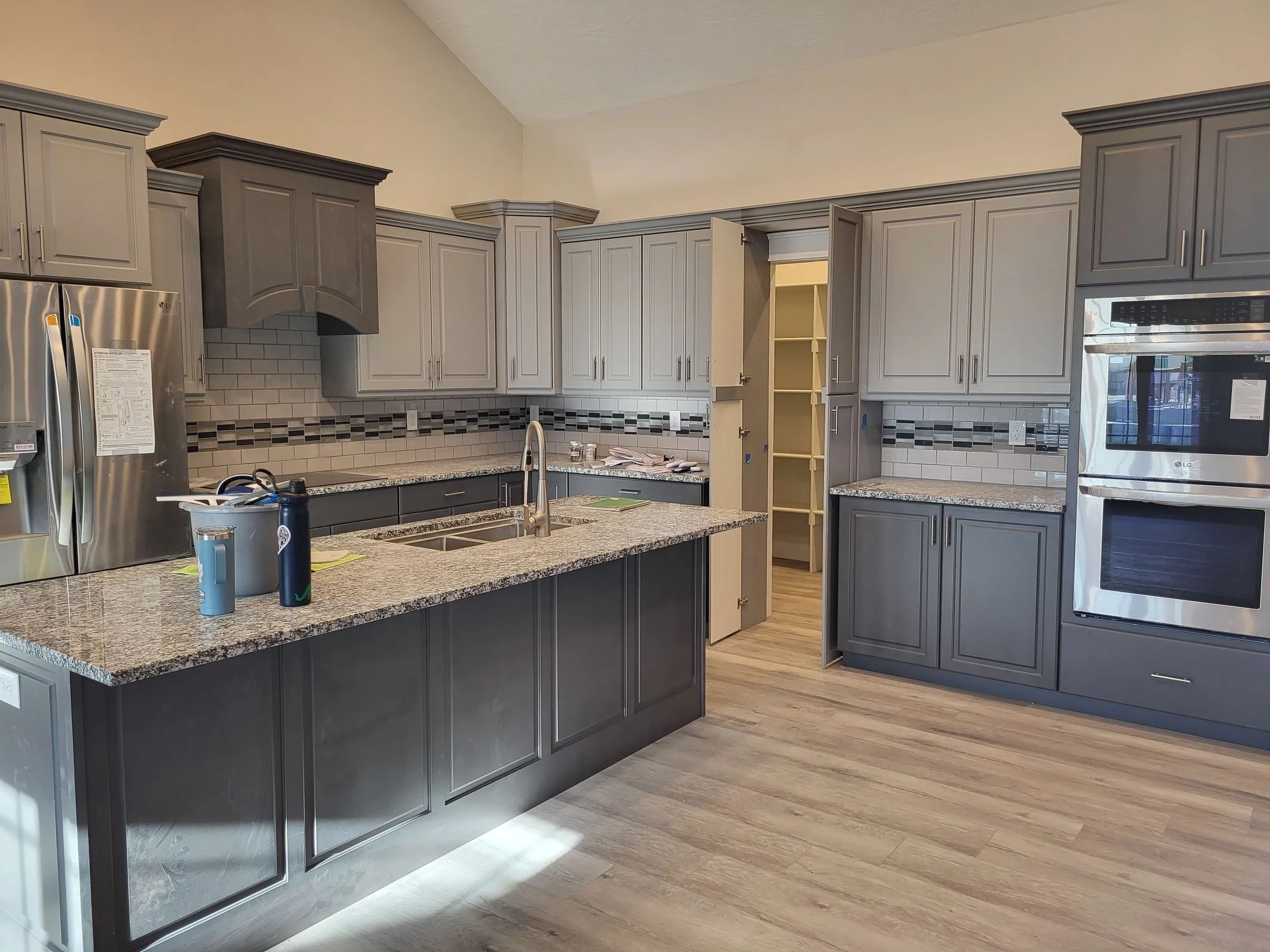 Modern kitchen with gray cabinets, granite countertops, stainless steel appliances, including a refrigerator and double oven, and a wood floor.