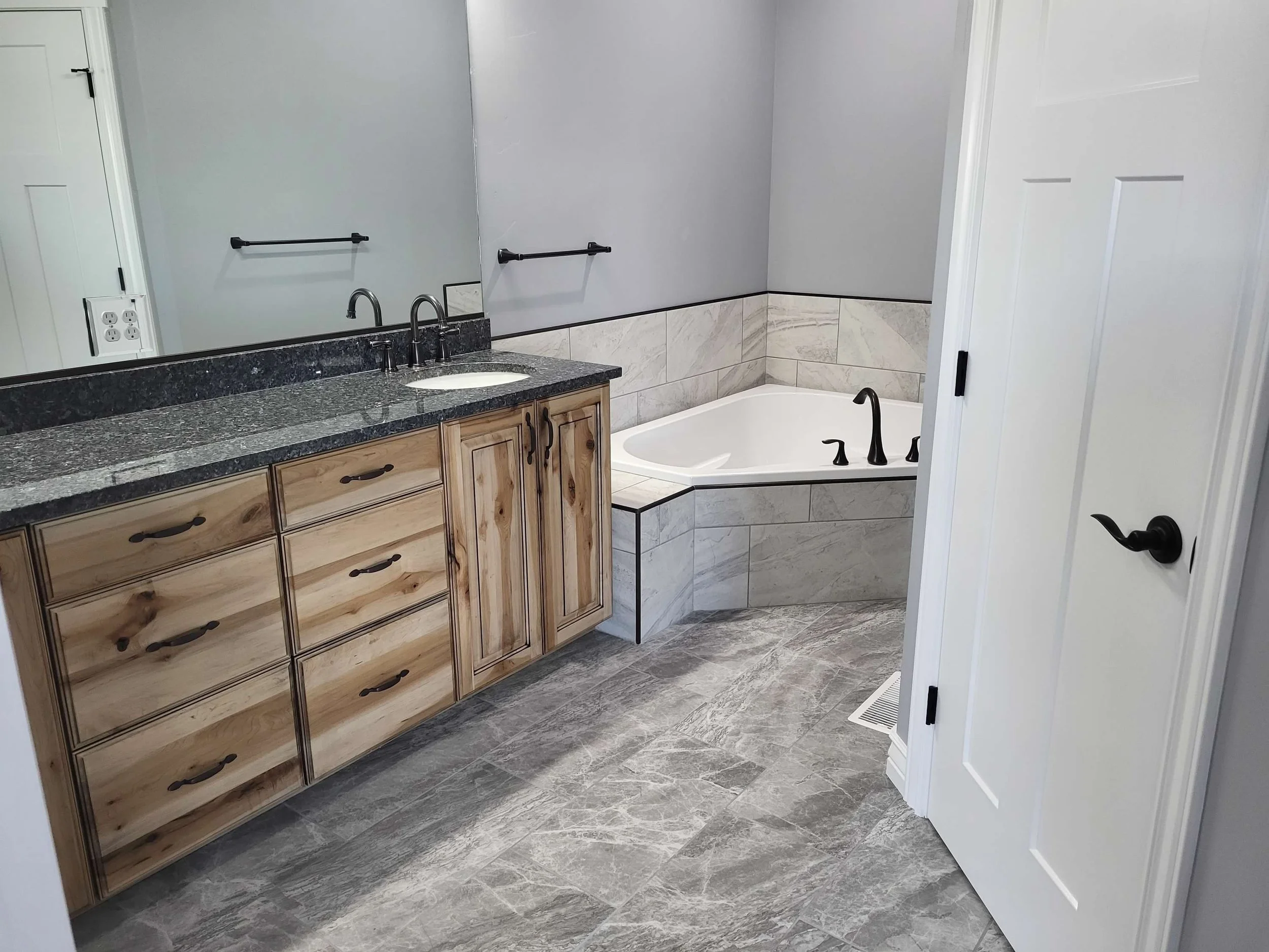 Bathroom with a granite countertop vanity cabinet, a large mirror, two black faucets, a corner bathtub with black fixtures, and grey tiled walls and floor.