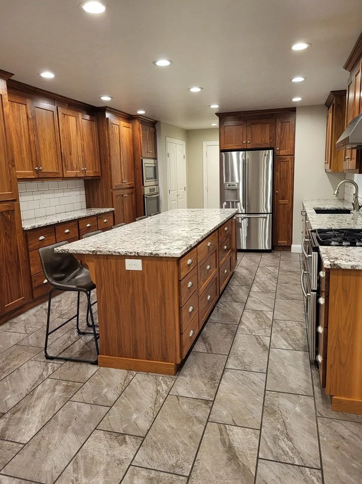 Modern kitchen with wooden cabinets, granite countertops, stainless steel appliances including a fridge, double oven, and stove, tiled floor, and a large kitchen island with drawers and a bar stool.
