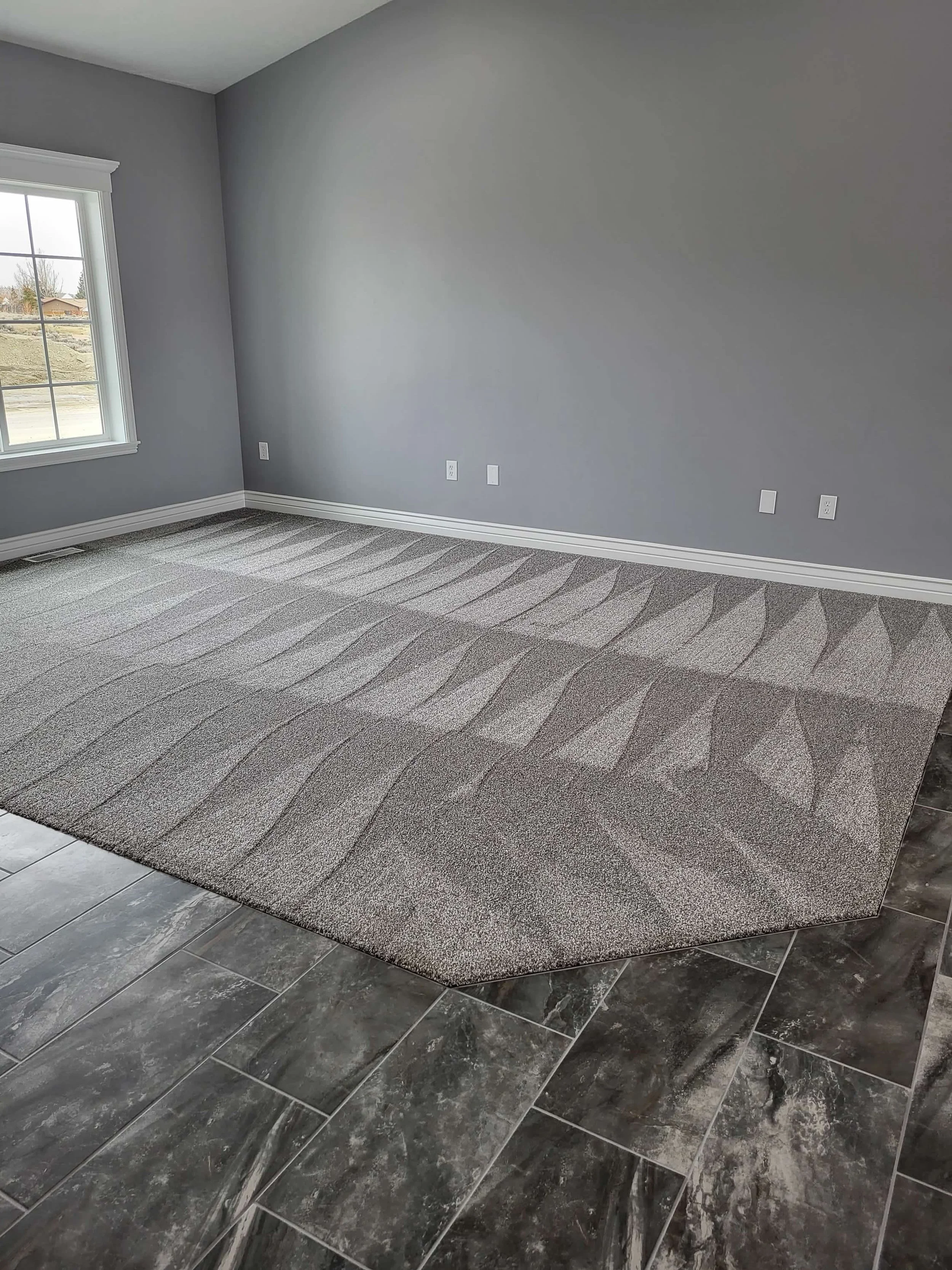 Empty room with gray walls, a large window with white trim, a carpeted floor with a pattern, and gray tiled flooring at the edge.