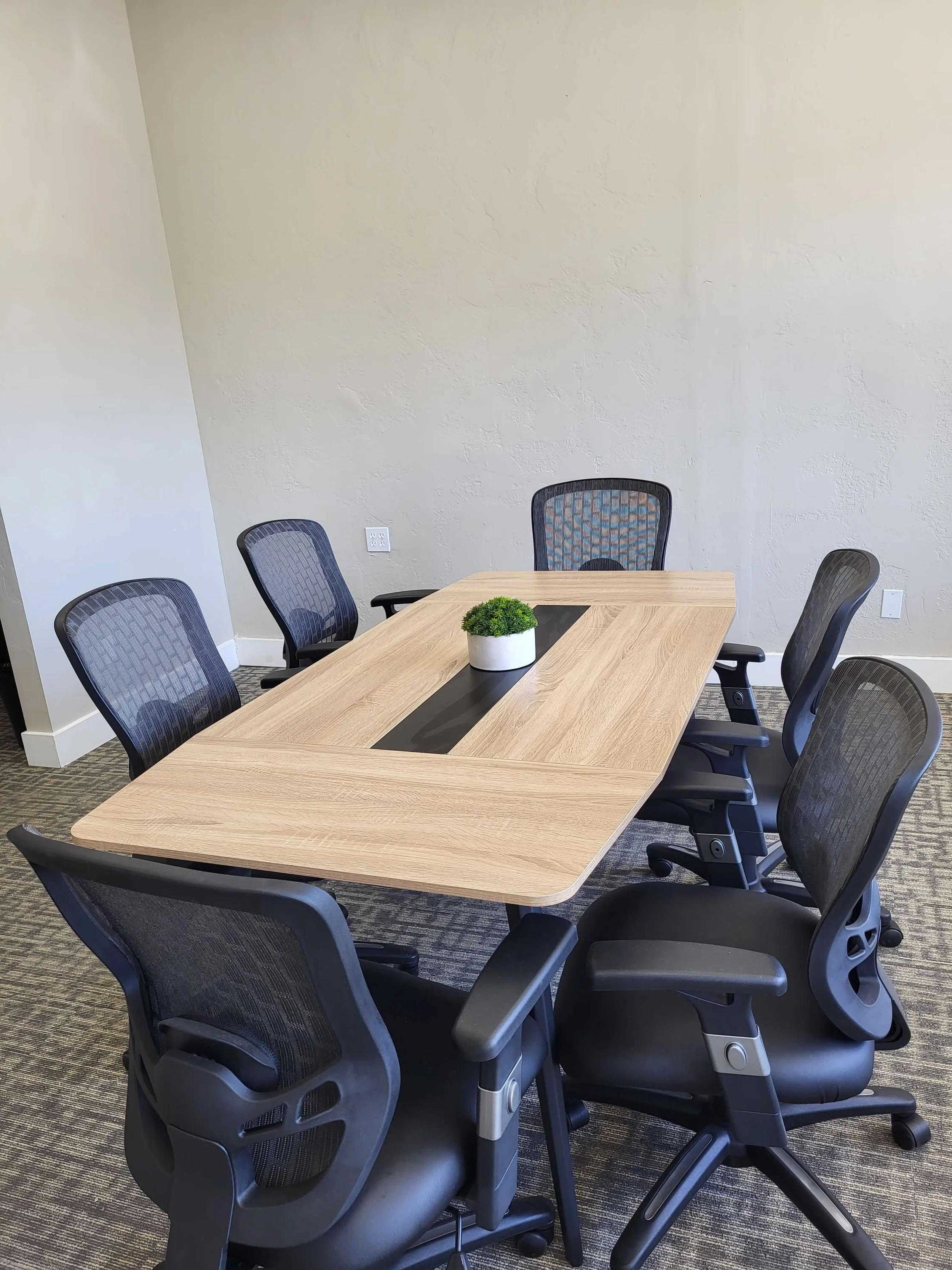 A modern conference room with six black office chairs around a wooden table. A small potted green plant is in the center of the table, and the room has plain light-colored walls and a carpeted floor.