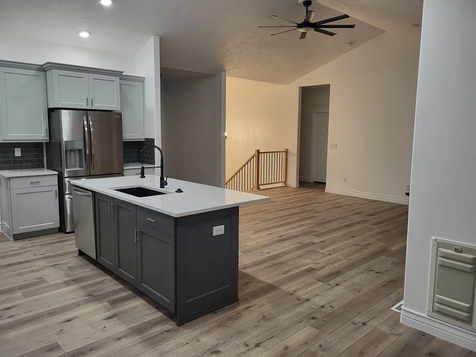 Open-concept kitchen with gray cabinets, white countertops, black backsplash, stainless steel refrigerator, and a kitchen island with a sink. Adjacent living space features a wood floor, ceiling fan, staircase with wooden railing, and a doorway.