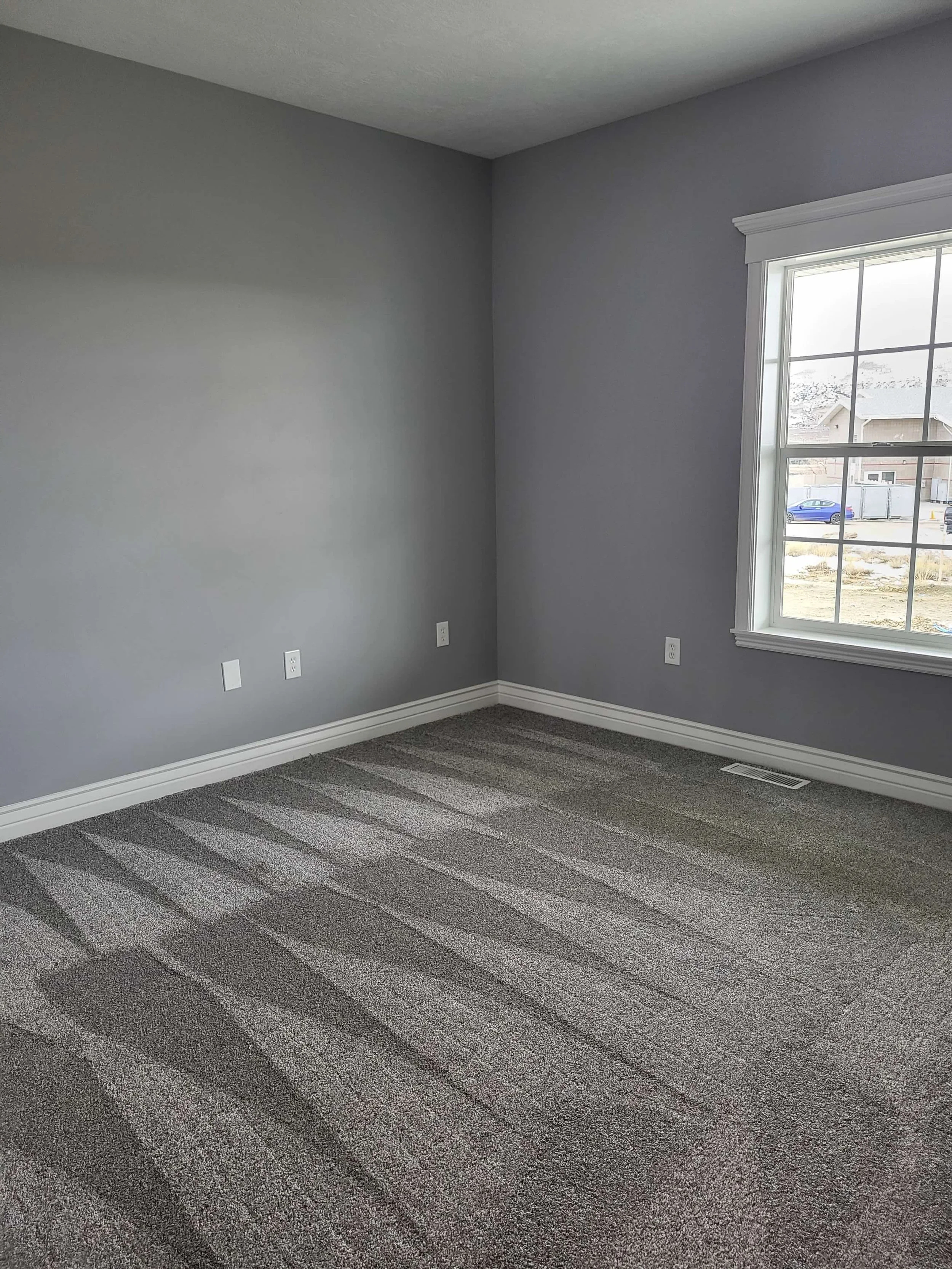 Empty room with gray walls, carpeted floor, and large window showing neighborhood outside.