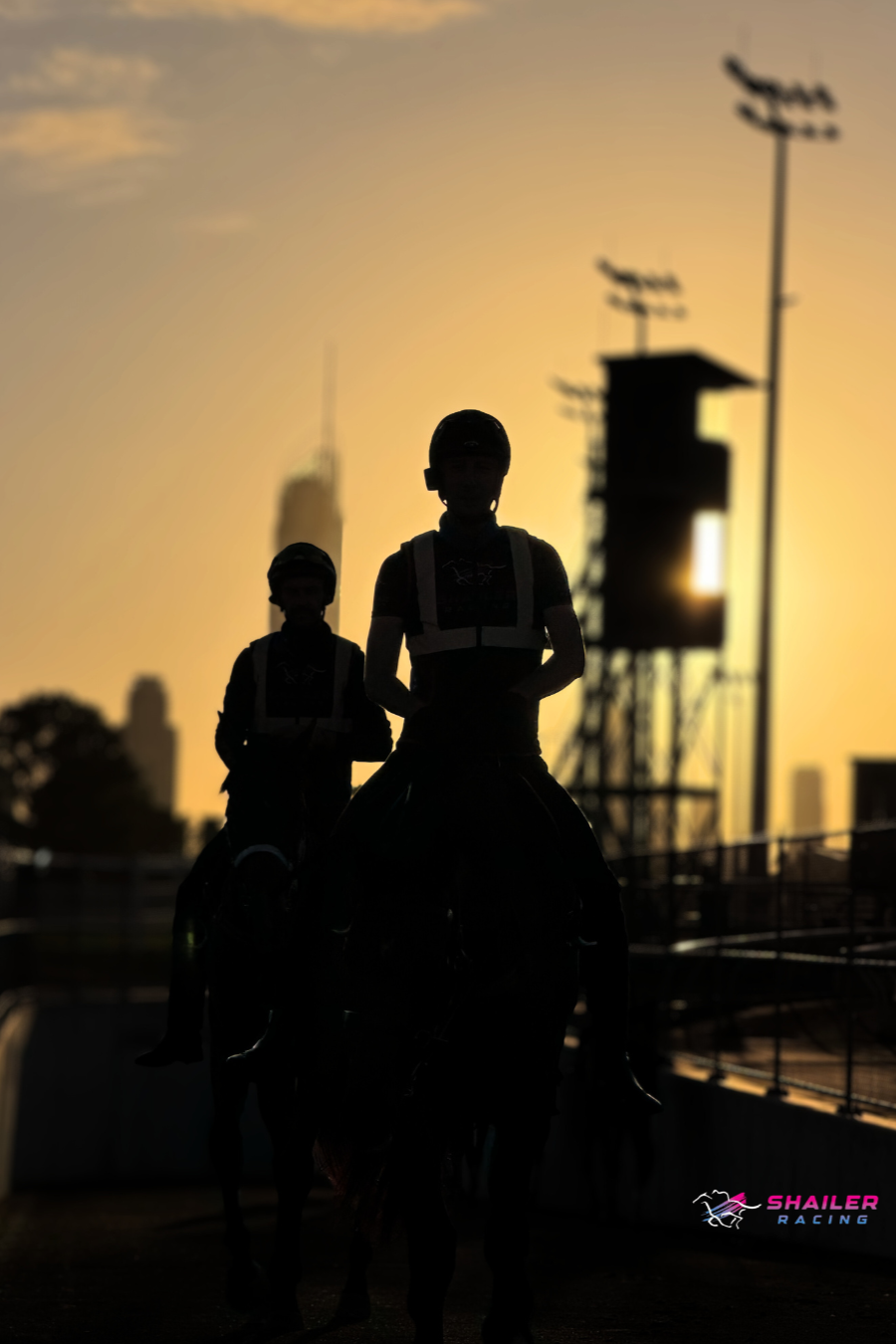 Silhouettes of two of Shailer Racings trackwork riders, wearing helmets and riding gear, against a gold coast sunrise sky at a racetrack with structures and light poles. Shailer Racing logo in the bottom right corner.