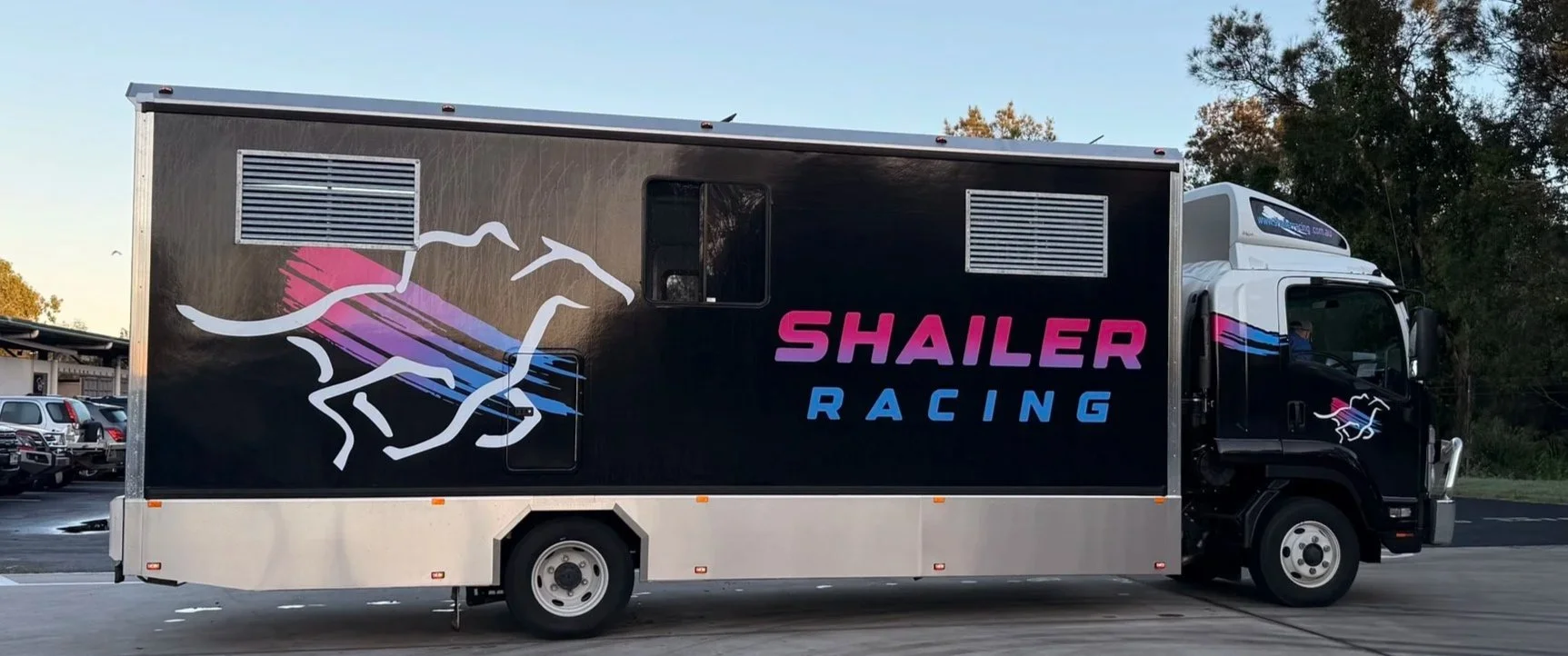 A racing truck with a black background, featuring a logo of four white horses and colorful lines, and the words 'SHAILER RACING' in pink and blue lettering.