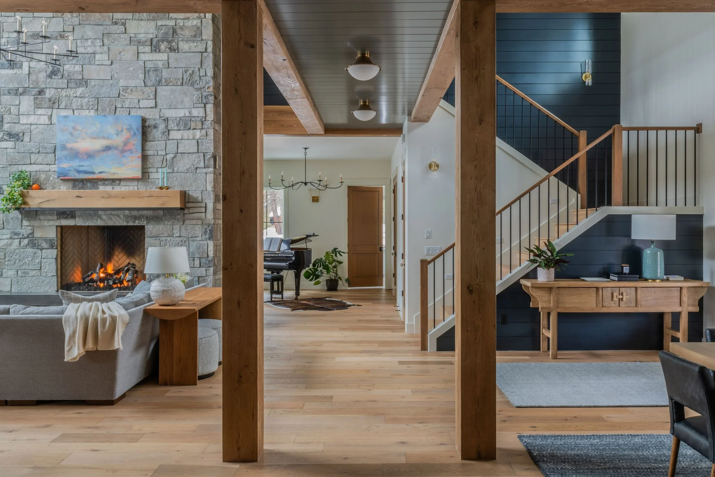 A spacious living area with a stone fireplace, a beige sofa, a wooden table, and a modern chandelier, leading to a hallway with hardwood floors and wooden accents, including a staircase and furniture.