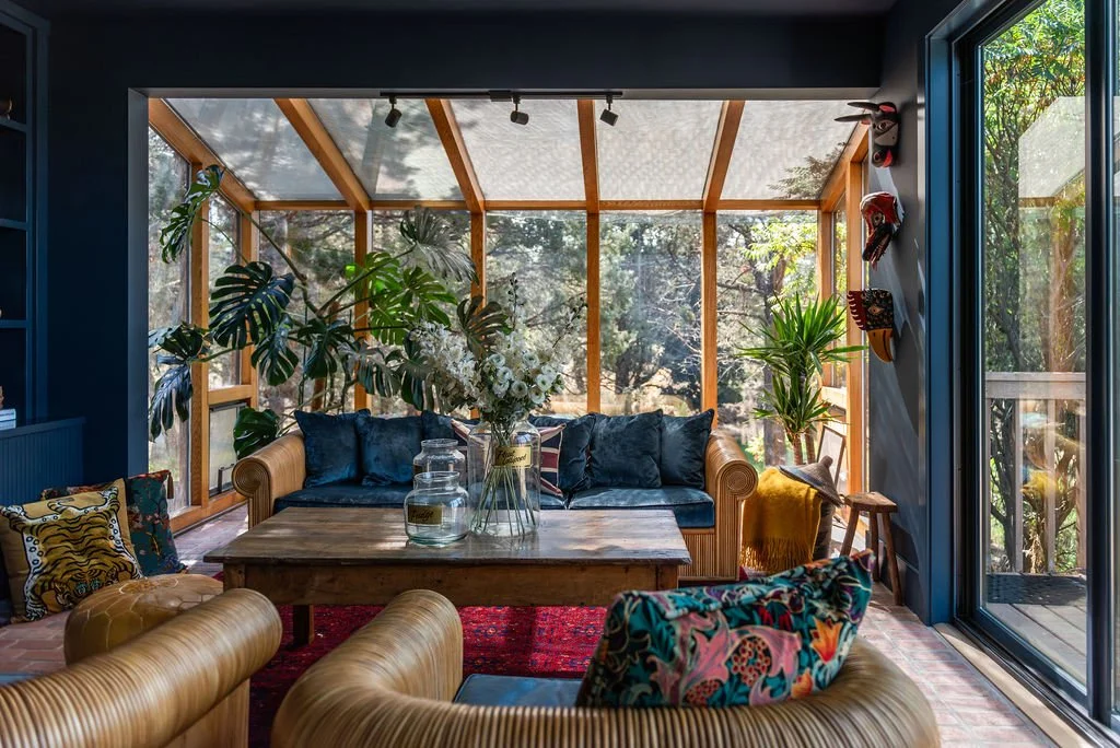 Sunlit indoor sunroom with tropical plants, blue velvet sofa, leather chairs, and wooden coffee table overlooking a forest