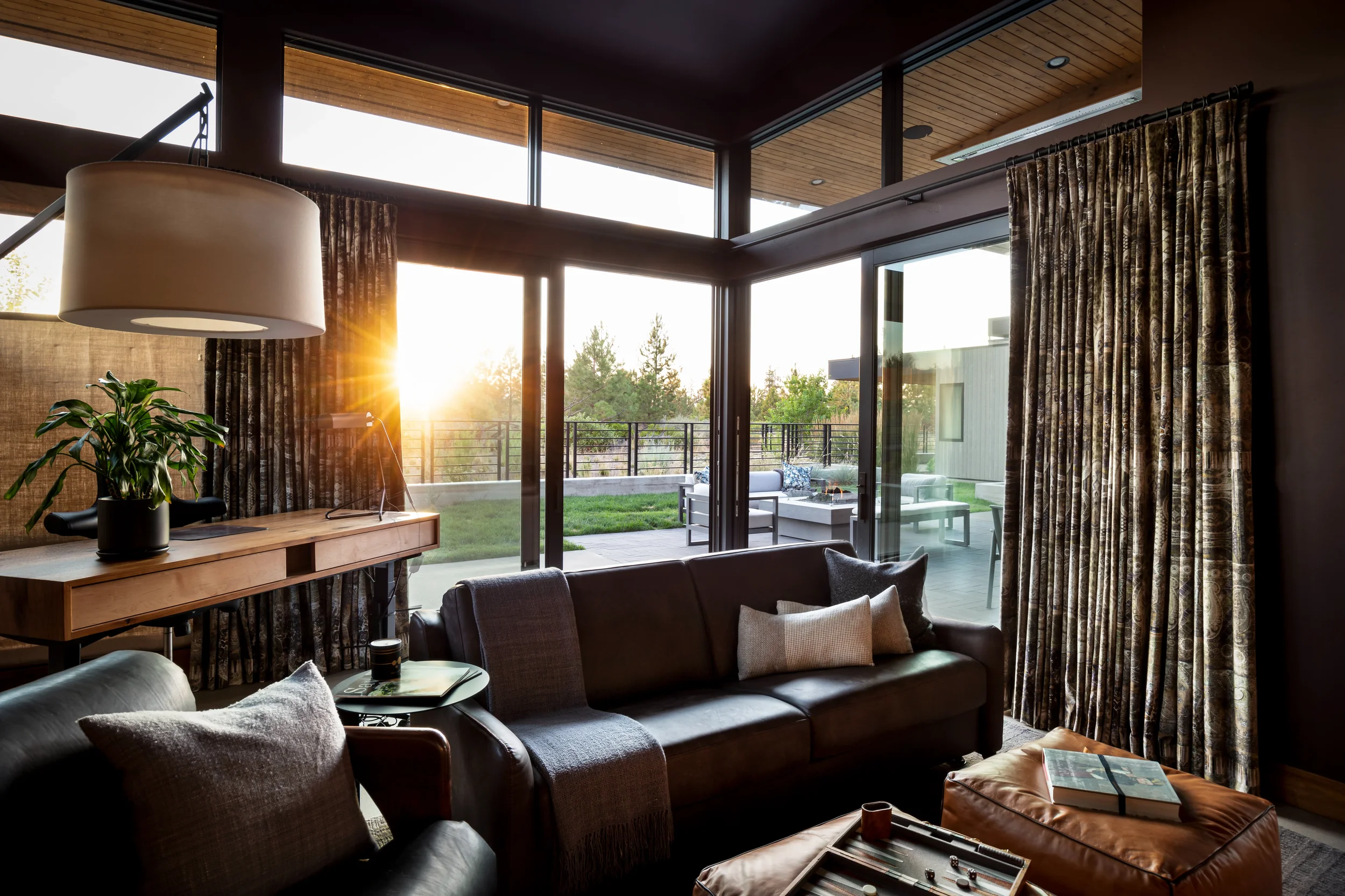 Modern living room with leather sofas, patterned curtains, floor-to-ceiling windows, and a sunlit outdoor patio