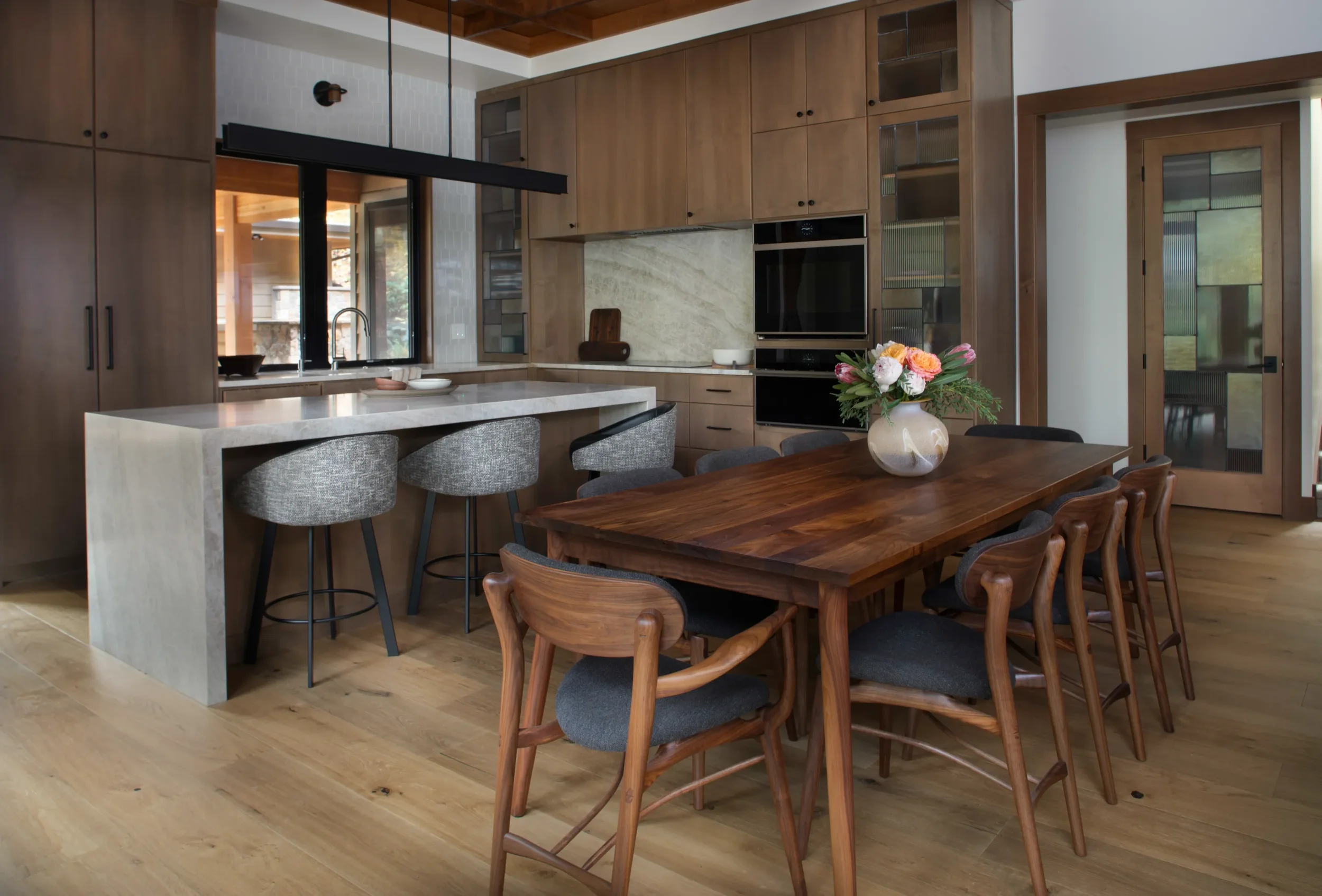 Modern wood-paneled kitchen and dining area with marble island, bar stools, built-in appliances, and a floral centerpiece on the dining table