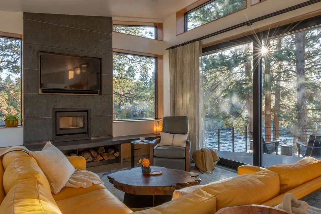 Modern living room with leather sofas, stone fireplace, wall-mounted TV, and large windows overlooking tall pine trees