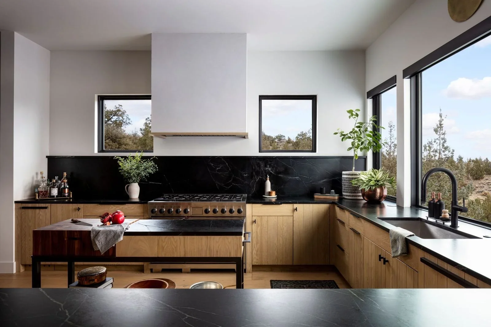 Modern kitchen with black marble countertops, wood cabinets, large windows, gas range, farmhouse sink, and potted plants