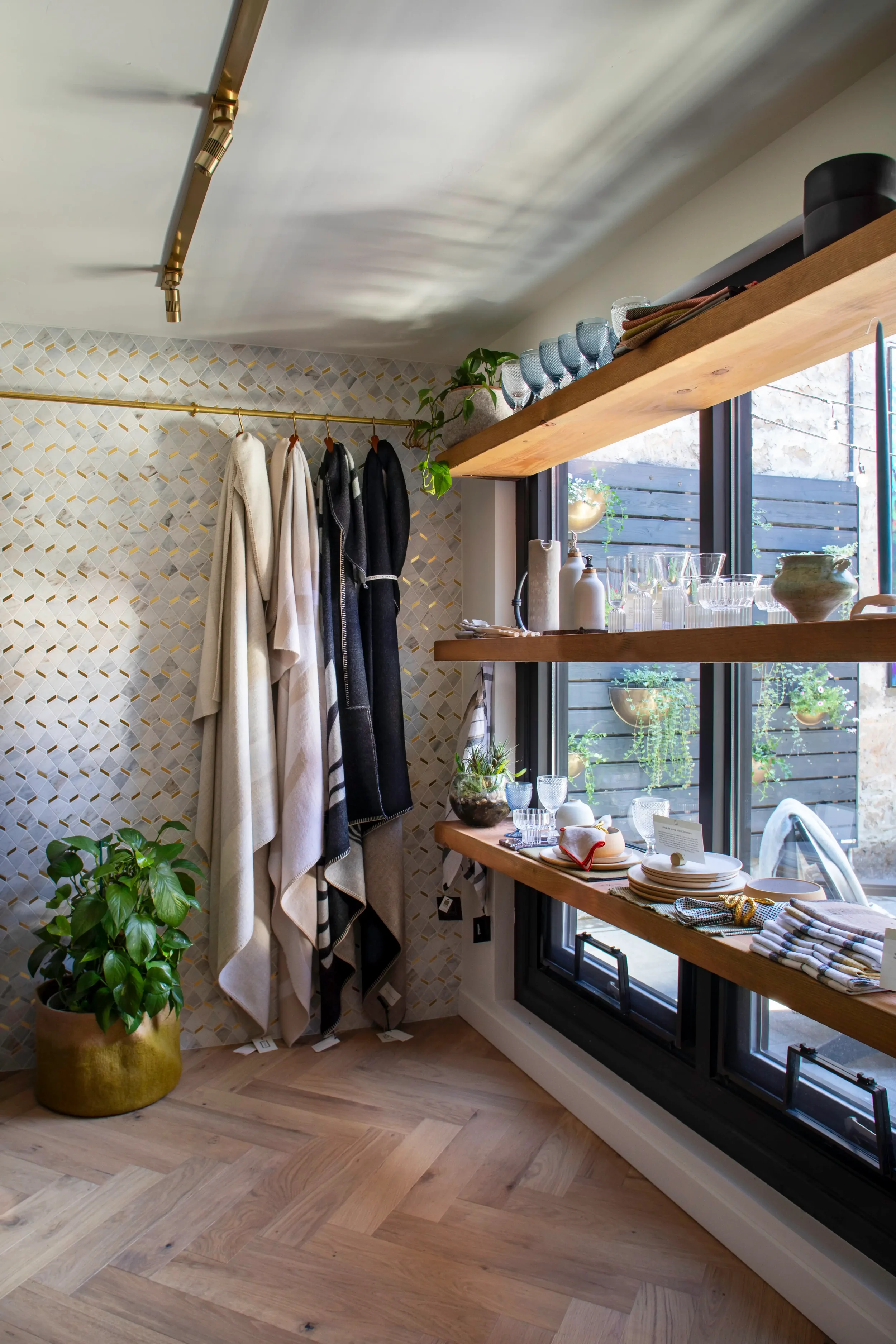 A cozy interior kitchen display with colorful glassware on a floating wooden shelf, hanging towels on a brass rod, a large leafy plant in a brass pot, window with potted plants outside, and a table set with plates, glasses, and utensils.