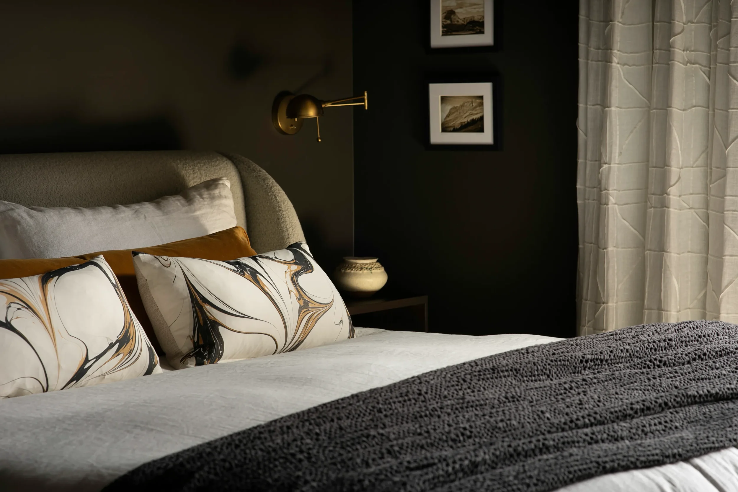 Elegant bedroom with patterned pillows, dark textured blanket, brass wall lamp, framed art, and white curtains