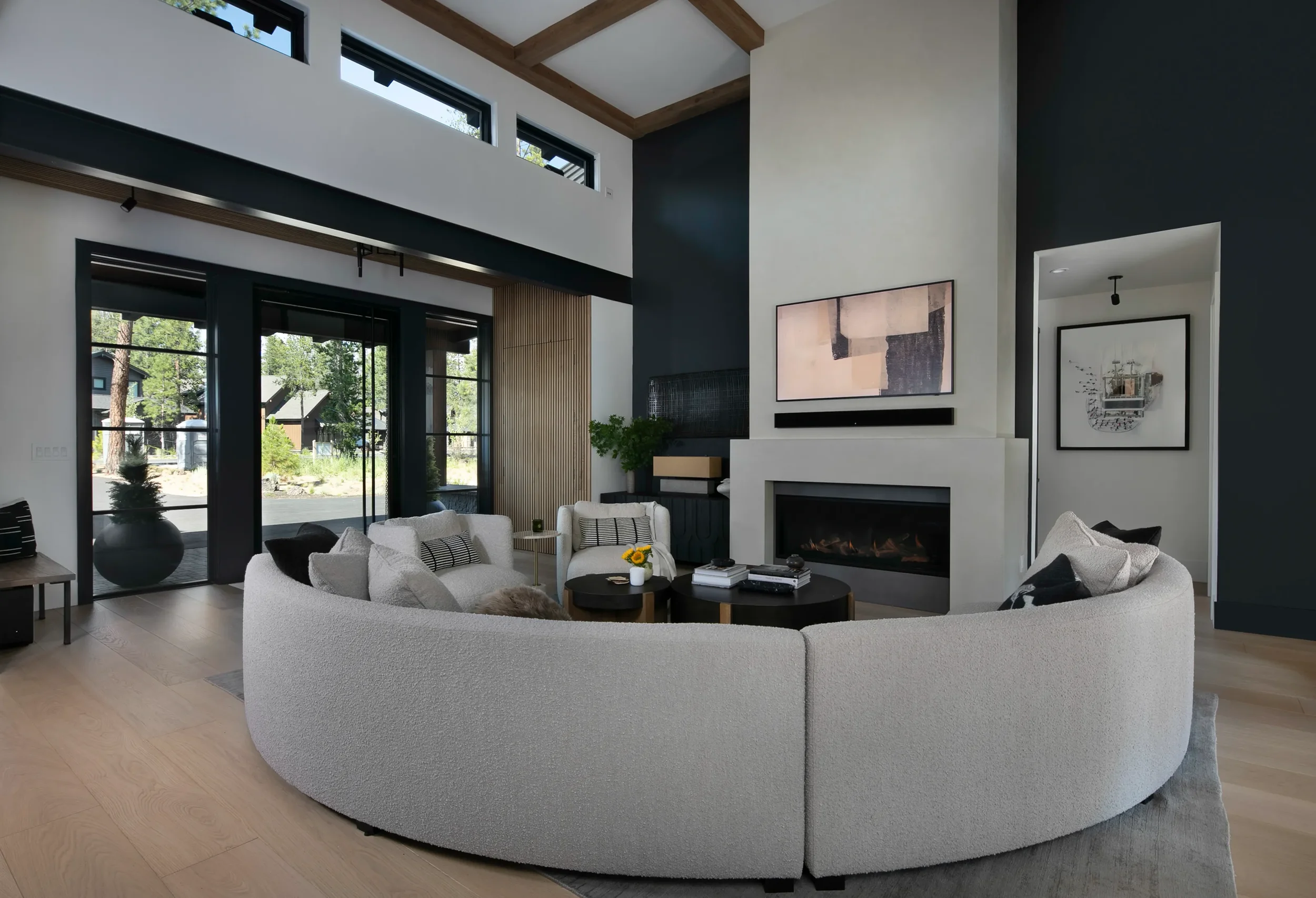 Modern living room with a curved sectional sofa, fireplace, abstract artwork, high windows, and exposed wood ceiling beams