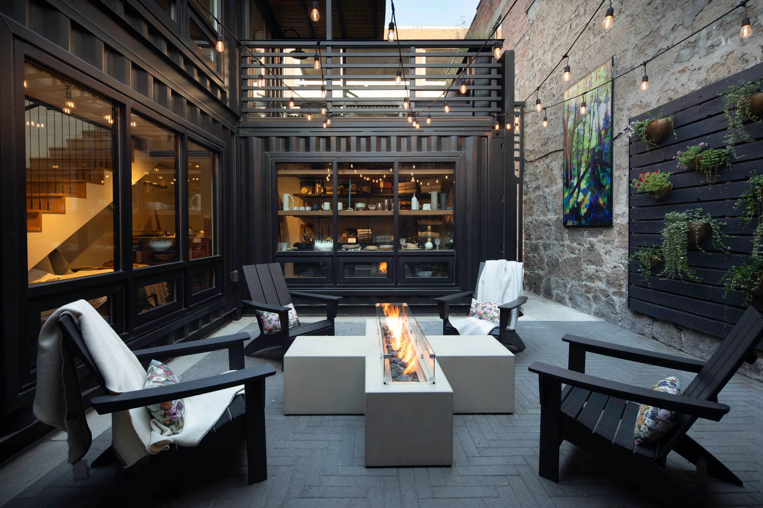 A modern urban outdoor patio with string lights, a central fire feature, and contemporary seating, enclosed by black metal and glass walls with a stairway and artwork on a stone wall.