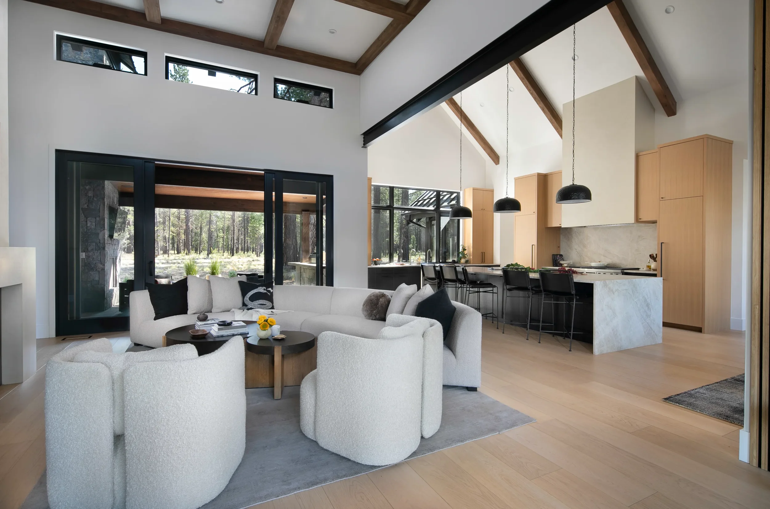 Modern open-concept living room and kitchen with vaulted wood-beam ceilings, white sofas, black pendant lights, and forest views